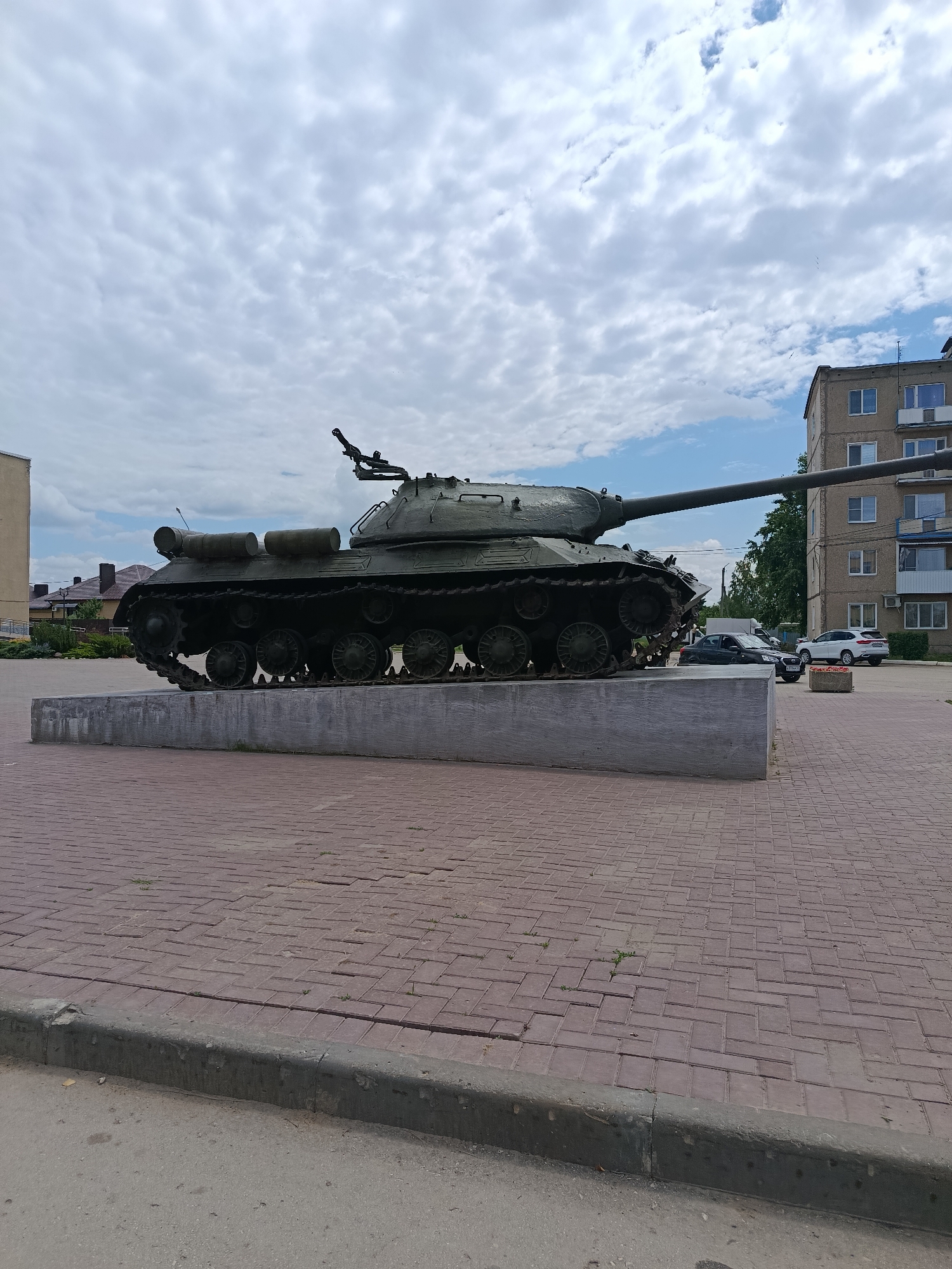 An IS-3 heavy tank is displayed as a monument on a concrete pedestal in the city of Marx, Russia. The tank, painted olive drab, is positioned on a paved square with red brick pavers. Behind the tank, a multi-story apartment building with light-colored walls and numerous windows is visible, along with trees and other buildings. The sky is overcast with scattered clouds. Several cars are parked near the apartment building. The foreground shows a concrete curb and asphalt road.
