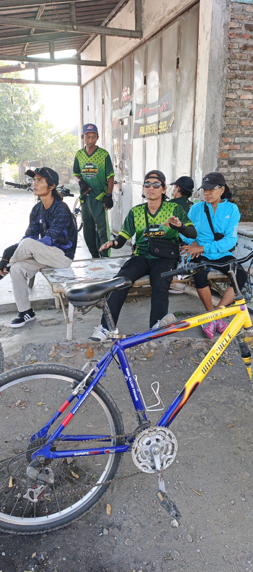Four individuals are gathered outdoors, seemingly taking a break. Two men in matching green and black cycling jerseys with the "FIN BIKE" logo are present. One is standing, while the other is seated and gesturing. A third individual, also in a green and black jersey, is standing near a metal garage door with various stickers, including "ALDER-92". A fourth person, wearing a dark blue jacket and a black baseball cap, is seated next to the gesturing cyclist.
A blue and yellow bicycle is prominently displayed in the foreground, with the words "ROADTECH-S" and "WIM CYCLE" visible on its frame. The bicycle has a water bottle cage and a Shimano derailleur. The individuals appear to be in the city of Kebonan, Indonesia, based on the presence of the "FIN BIKE" cycling group, which is associated with the region. The environment is dusty and appears to be an outdoor gathering area, possibly near a business or workshop, with a weathered garage door in the background. The lighting suggests it is daytime, with sunlight casting shadows.