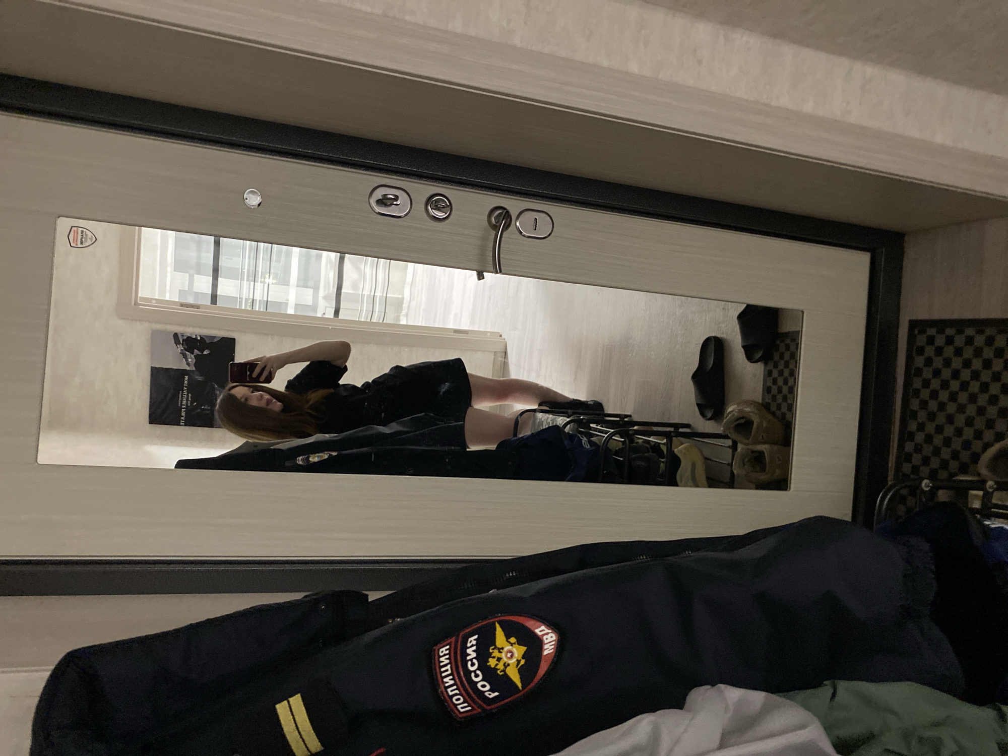 The image shows a mirror reflection of a young woman with long brown hair, lying down and taking a selfie with her phone. She is wearing a dark shirt and shorts. Behind her in the reflection is a window with curtains, and a poster on the wall. Shoes and clothing are scattered on the floor. In the foreground, a dark blue jacket with a patch in Russian is visible. The patch reads "РОССИЯ ПОЛИЦИЯ МВД". The setting appears to be an indoor space, possibly an apartment or dormitory room. There is an assumption of the location due to the patch on the jacket stating Russian police. A forensic analysis of the image cannot reliably pinpoint a location; however, based on the presumption of a police jacket, Chaikovsky, Russia, is assumed.