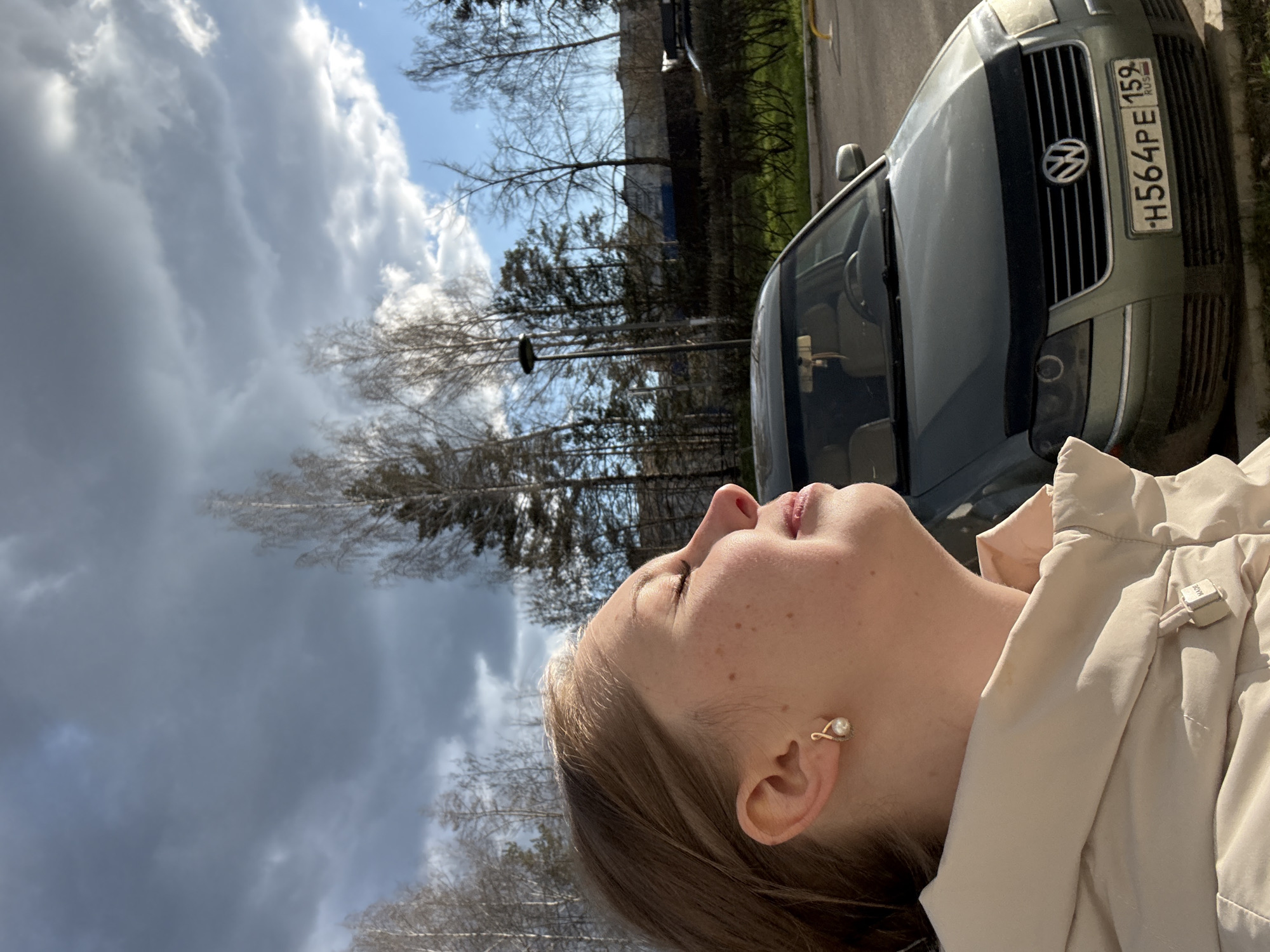 The image, rotated 90 degrees clockwise, prominently features a young person in profile in the foreground, looking upwards with eyes closed or squinting. She has light brown hair and fair skin with freckles, and wears a light-colored jacket. A distinctive gold-toned ear cuff with a pearl-like adornment is visible on her ear. A small, rectangular clip-on device, possibly a microphone, with "MADE" partially legible, is attached to her jacket collar.
In the mid-ground, a dark green or grey Volkswagen car is parked, with its front grille, emblem, and headlights visible. The car bears a Russian license plate.
The background shows several bare trees, suggesting a late winter or early spring setting. The sky above is a mix of bright blue patches and large, textured grey and white clouds, indicating a partly cloudy day.
The activity depicted is the person gazing upwards, seemingly enjoying the light or lost in thought. The time of day is daytime, with visible sunlight. The license plate "H564PE 159 RUS" identifies the location as Russia, specifically referencing the 159 region (Perm Krai).