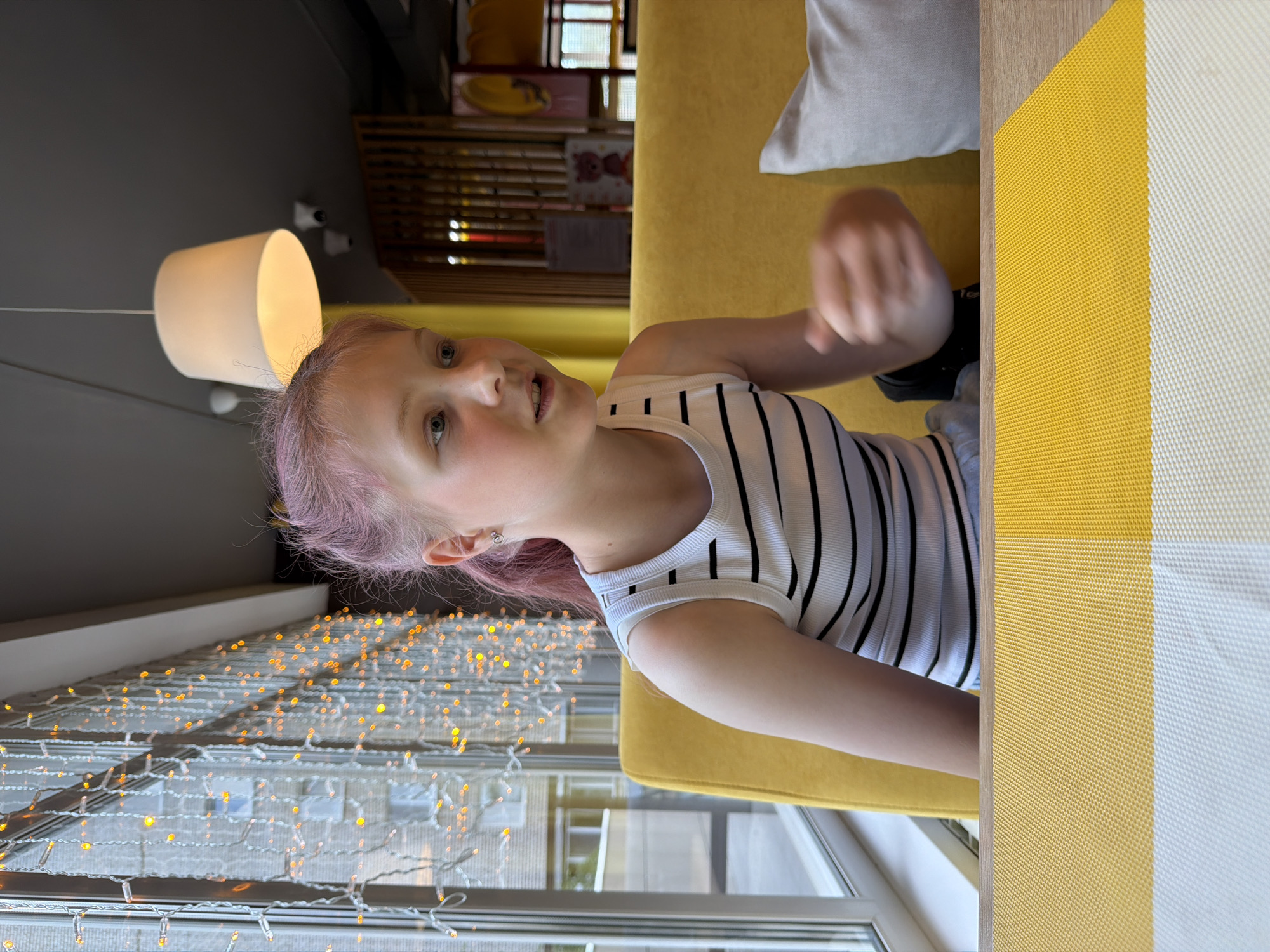 An image shows a young Caucasian girl with pink hair sitting on a yellow sofa in a restaurant. She is wearing a black and white striped tank top and light blue shorts. Her eyes are blue and she is looking to the right of the frame. Her right hand is blurred as if she is in motion. The background of the image shows a large window with string lights hanging across it, a hanging lamp, and wooden partitions. The girl is sitting at a table with a yellow and white placemat. The city of Chaikovsky, Russia is not visible in the image.