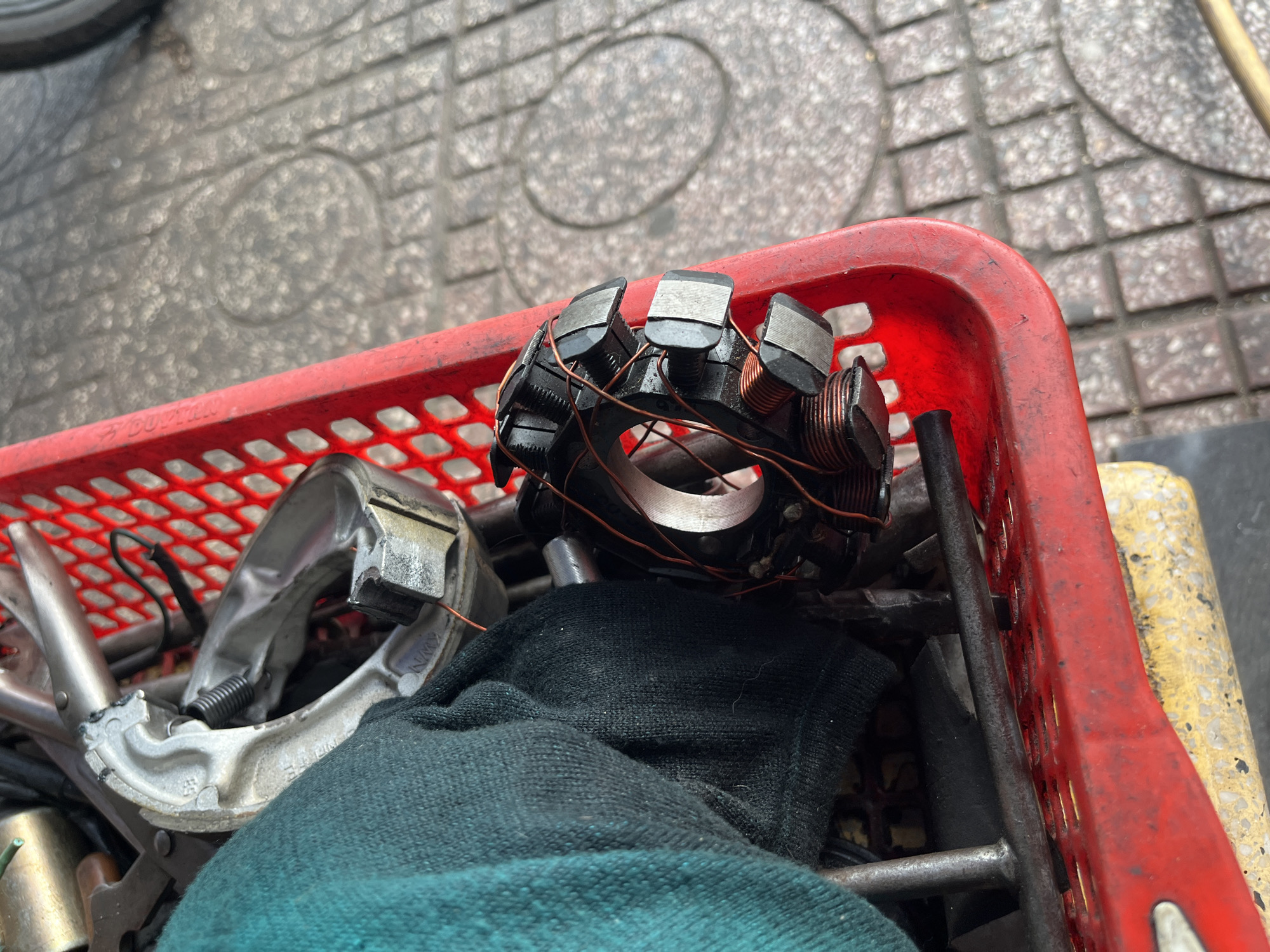 A close-up captures a red plastic basket containing mechanical components. Dominating the view is a motorcycle stator, its copper windings and metal laminations prominently displayed. Alongside it are various tools and a silver brake shoe assembly, partially obscured by a dark-colored glove or cloth. The basket sits on what appears to be a concrete surface with a patterned design. While I am unable to confirm the specific location, the information provided states the image is in Bac Nha Trang Ward, Vietnam. The scene suggests a motorcycle repair or maintenance context.