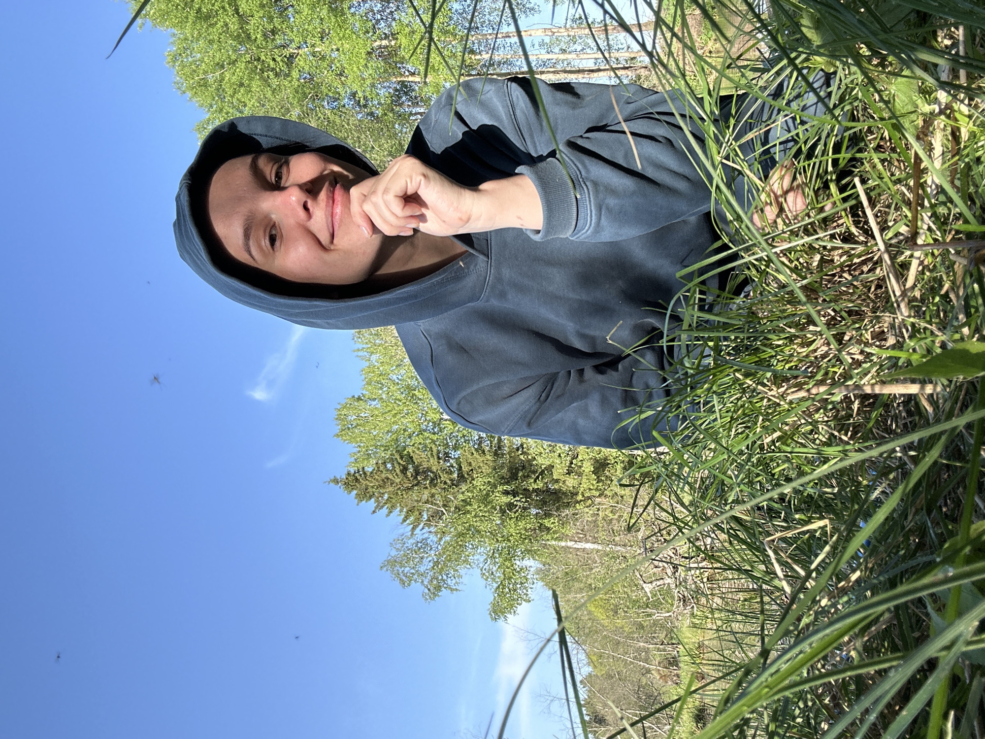A female in a blue hooded sweatshirt is visible from the chest up, with a relaxed posture and smiling while resting her hand on her chin. She is likely outdoors, surrounded by tall green grass and under a clear blue sky with a few sparse clouds. Background elements suggest a natural setting with trees. No specific identifying details place the scene within Bormist, Russia.