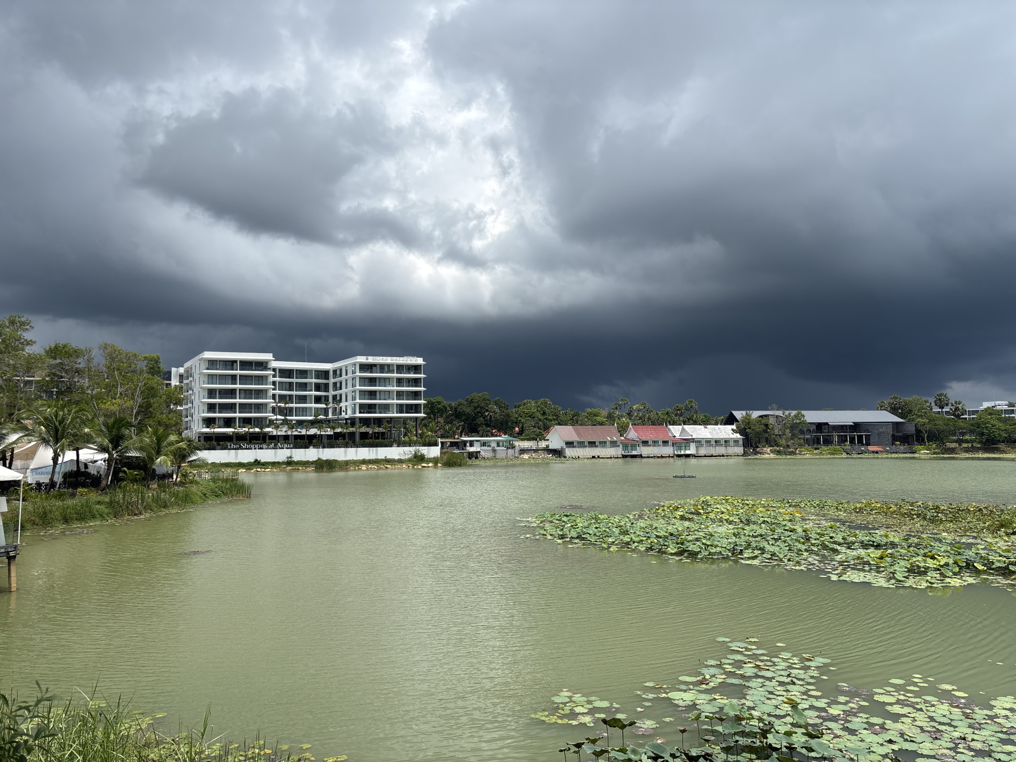 The image captures a view of a lake in Choeng Thale, Thailand, under a stormy sky. A modern white building with multiple floors is visible on the left bank of the lake, near some palm trees and other vegetation. Several smaller buildings with red and grey roofs line the opposite shore. The lake is covered with lily pads and other aquatic plants. The sky is dominated by dark, ominous storm clouds.