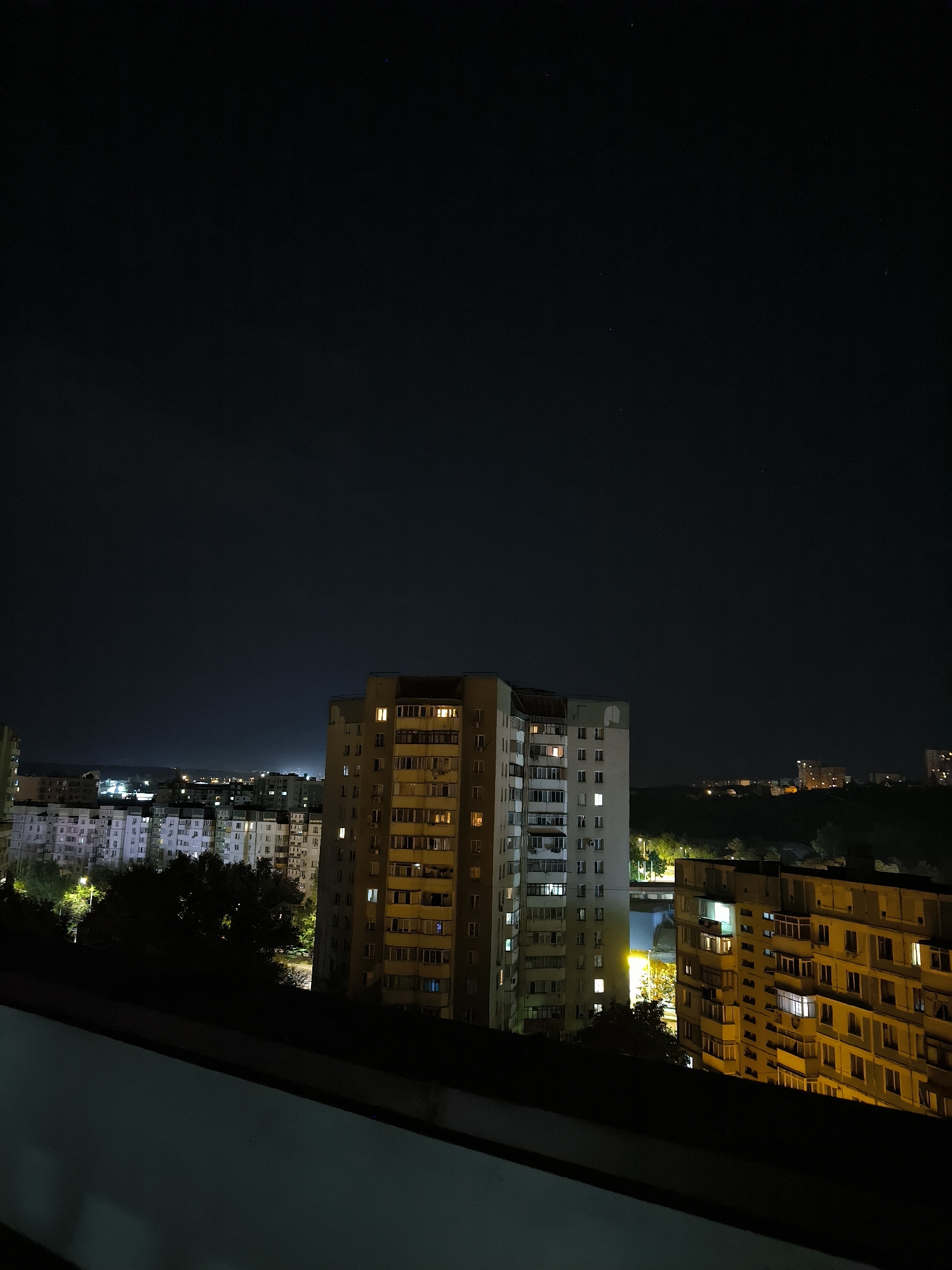 The image depicts a nighttime cityscape in Chișinău, Moldova, viewed from a high vantage point, possibly a balcony or rooftop. The foreground is dominated by a dark, blurred railing or ledge. In the middle ground, several apartment buildings are visible, illuminated by the lights emanating from their windows. The building on the right, a yellow-toned structure, has numerous windows, many of which are lit, suggesting occupancy. To its left, a taller, multi-story apartment building with a white and grey facade also shows illuminated windows. Further in the background, rows of similar apartment buildings stretch across the horizon, their lights creating a scattered pattern against the dark sky. Some trees are visible between the buildings, their shapes softened by the darkness and illuminated by streetlights. The sky above is a deep, dark blue, with a few faint stars visible. The overall impression is that of a residential area at night, with the city's lights painting a picture of urban life.