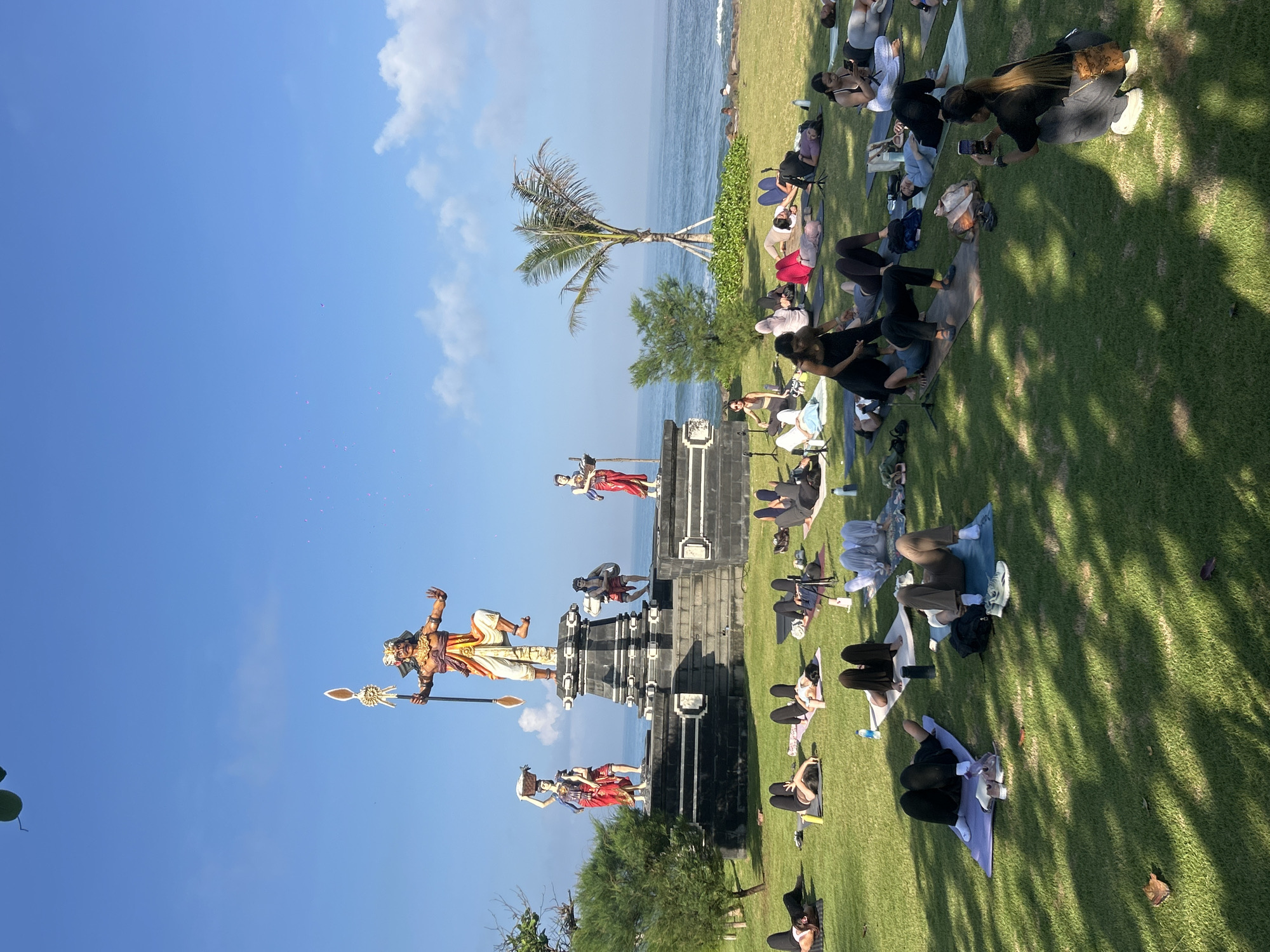 A wide-angle shot captures a scene in Mengwi, Indonesia. The location appears to be a grassy area near the coastline, with the ocean visible in the background. A statue of a figure holding a spear stands prominently, with other statues visible on the tiered structure. People are lying on yoga mats in the grass, several are using their phones, possibly in a yoga class or recreational activity. Shadows are cast on the grass from trees or other structures. A palm tree is near the beach. The sky is bright blue with some white clouds.