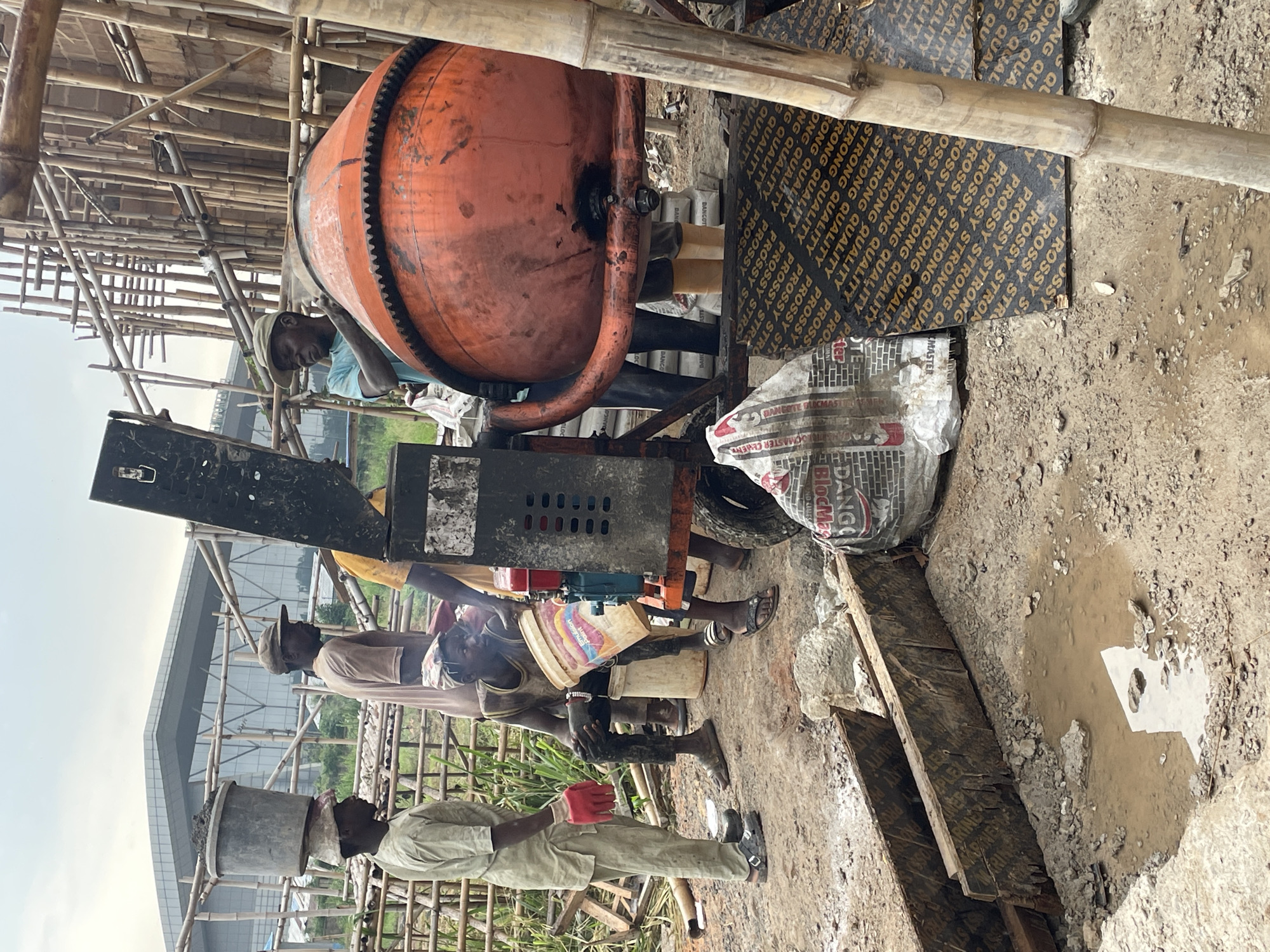 In Okinano, Nigeria, a construction site is captured under an overcast sky. Four individuals are present, engaged in activities related to a cement mixer. One man, wearing a light-colored outfit and red gloves, carries a large bucket of what appears to be cement on his head. Another man, in a light brown shirt and a cap, stands near the mixer. A third man, in a blue shirt, is seen interacting with the large orange cement mixer. The fourth person, wearing a sleeveless shirt and dark pants, sits on a white bucket holding another bucket. The cement mixer is positioned on a wheeled platform. The ground is muddy and uneven, with scattered debris, wooden planks, and a puddle of water. In the background, bamboo scaffolding surrounds a partially constructed building, and a modern building with glass windows is visible in the distance. The scene conveys a sense of ongoing construction work in a developing area.