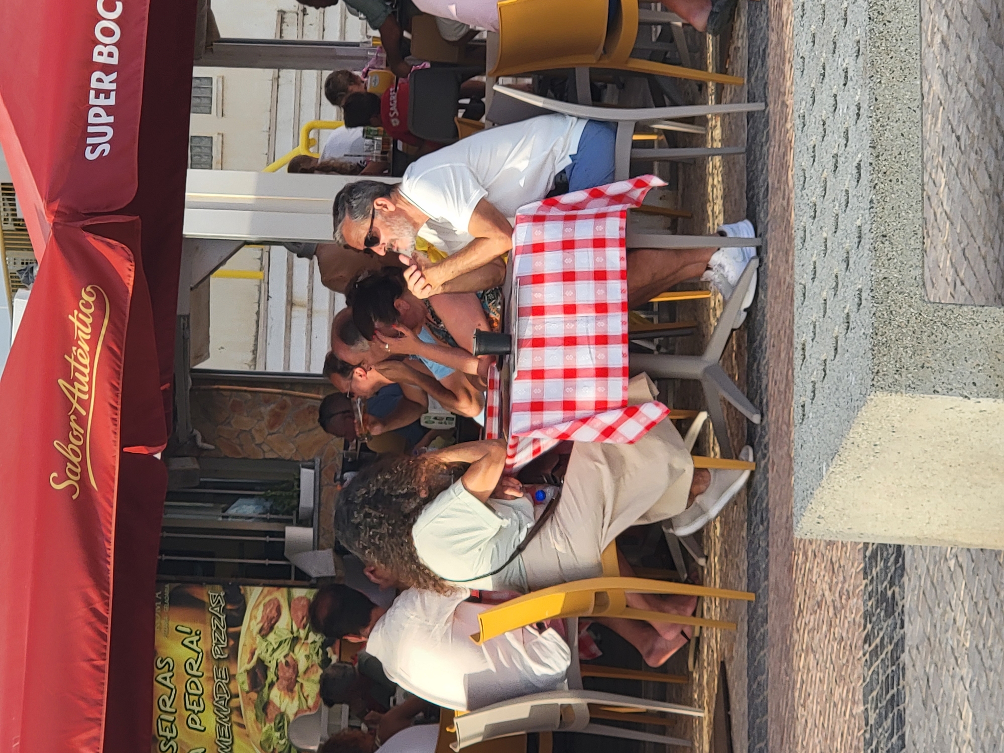 An outdoor seating area of a restaurant with red umbrellas is depicted in Quarteira, Portugal. A couple is seated at a table covered with a red and white checkered tablecloth. The man, wearing a white polo shirt and blue shorts, has his hands clasped in front of him and is looking to the side. The woman, with curly dark hair, is wearing a light-colored top and a long skirt, leaning on her hand and looking down. Other patrons and tables are visible in the background. The ground is paved with patterned stone. A sign on the umbrella reads "SUPER BO."