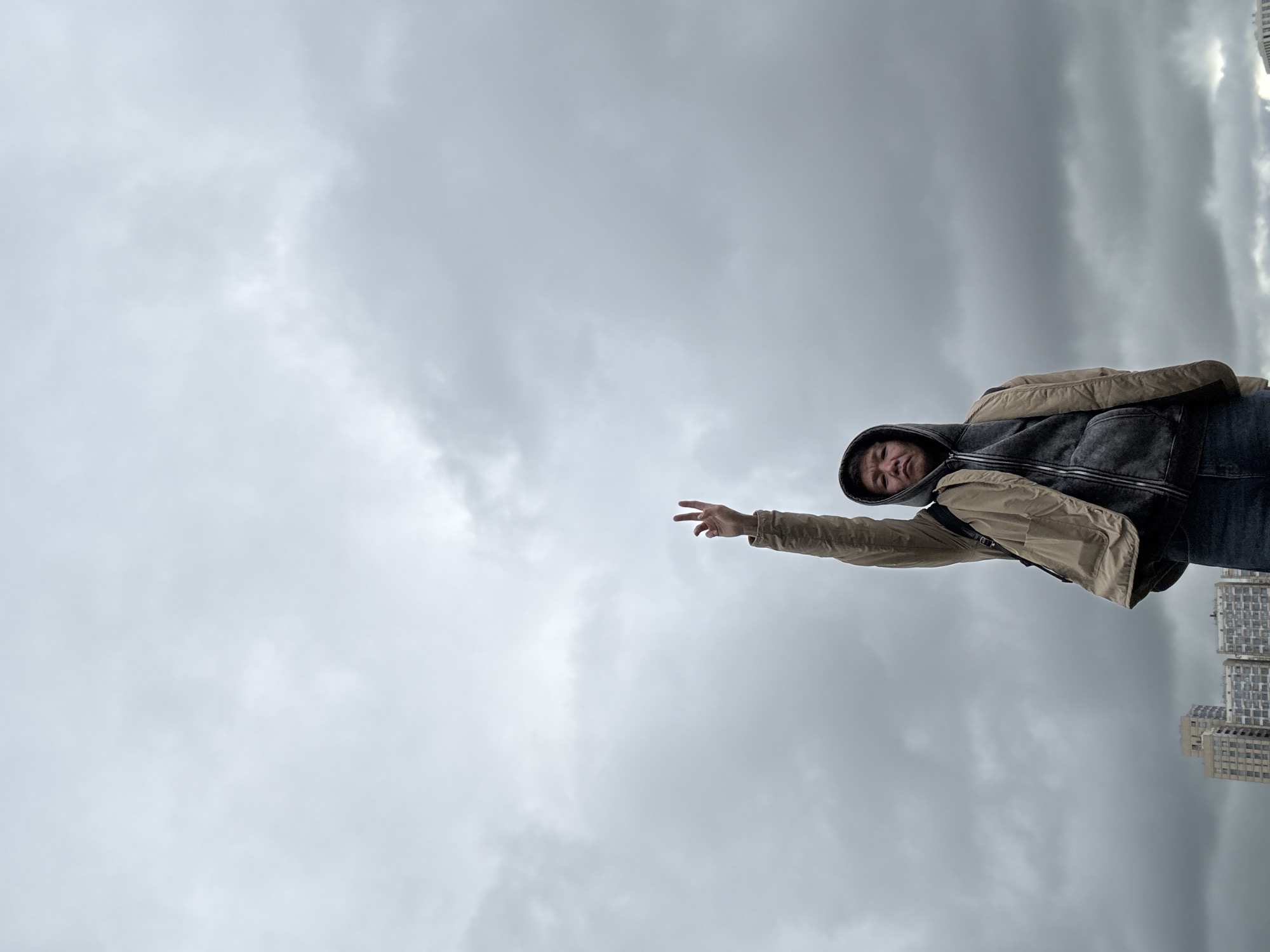 A man wearing a hooded jacket is reaching out towards the left side of the frame. He appears to be standing in Khimki, Russia, as multi-story buildings are visible in the lower right corner of the image. The sky is overcast with grey clouds.