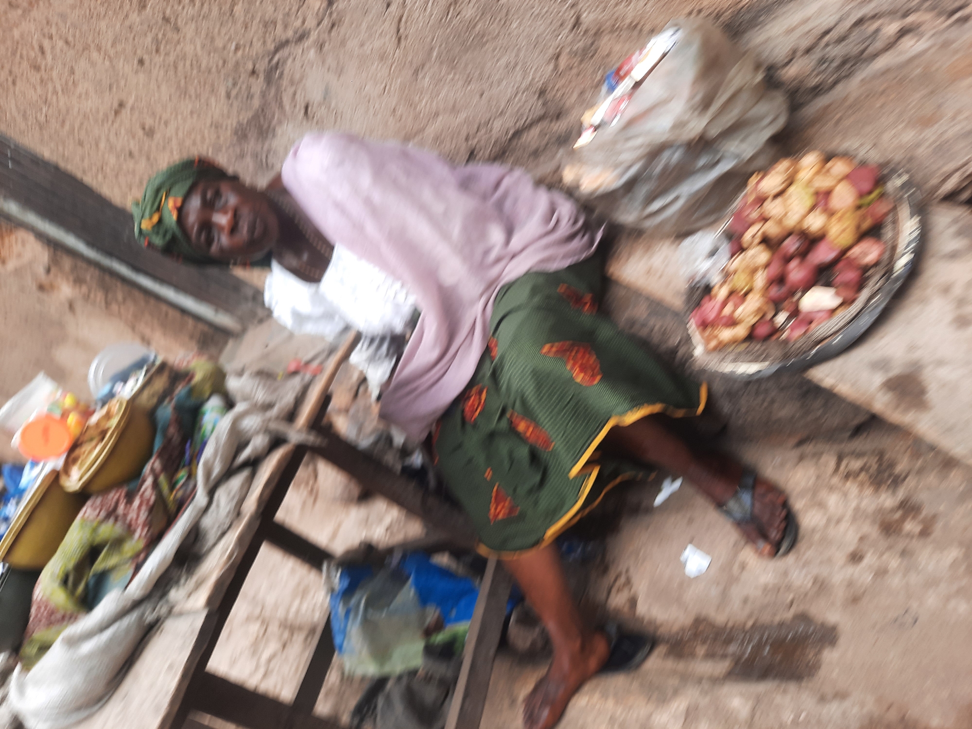An eye-level shot shows an older woman lying on a makeshift bed or structure in what appears to be an outdoor or semi-outdoor market setting in Jos, Nigeria. The woman, who has dark skin, wears a green head wrap, a white top, a pink shawl, and a green and yellow patterned skirt. She lies on her side, appearing to rest.
Next to the woman, on the right side of the image, is a pile of produce, including mangoes or similar fruits in a tray. There is also a white plastic bag or container next to the produce. A yellow bowl or container filled with items is on a wooden structure or stand to the left. The area appears to be a trading area with a concrete floor and a wall in the background. The scene has a slightly disheveled or worn appearance.