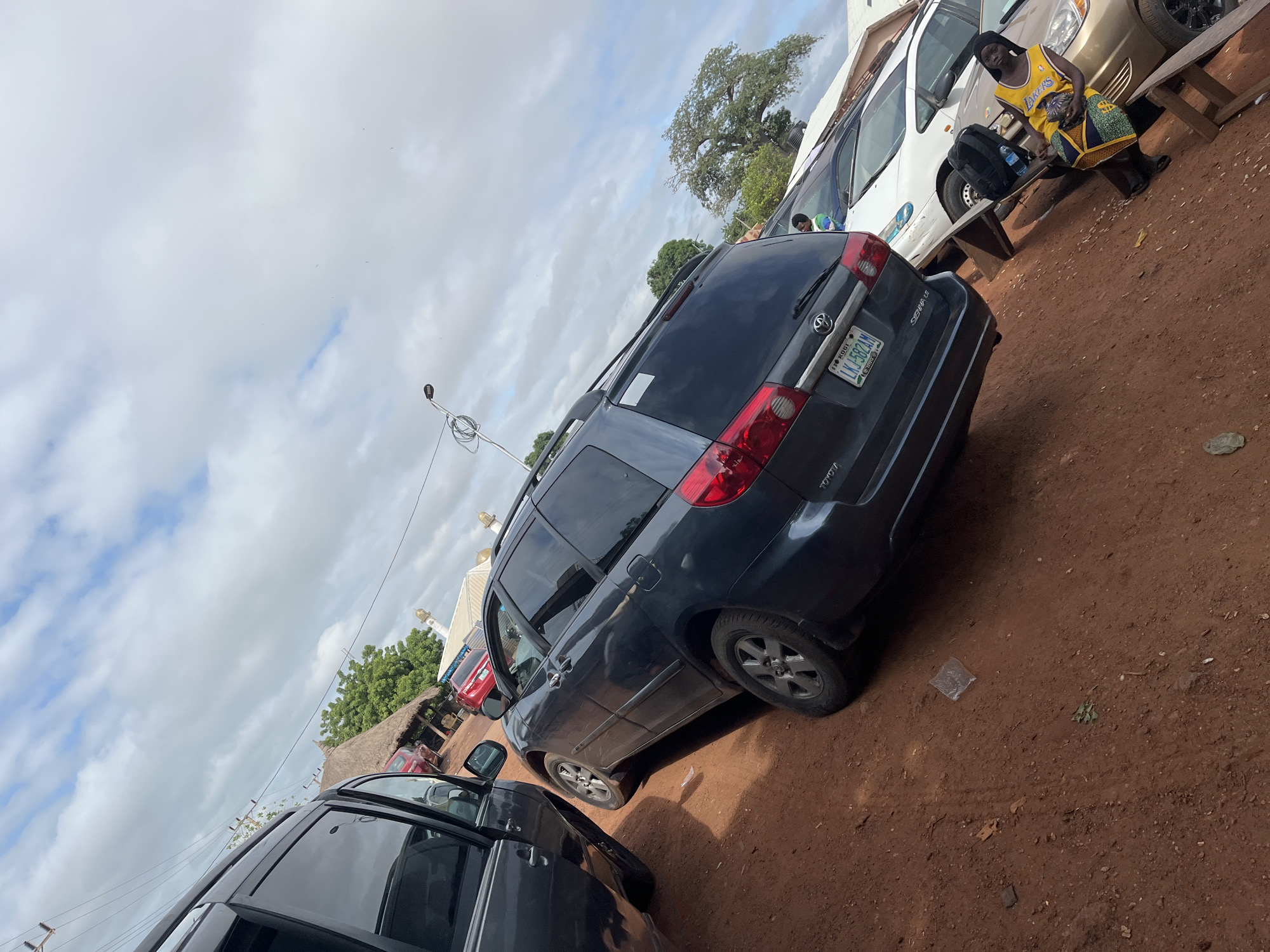 The image depicts an outdoor scene in Idah, Kogi State, Nigeria, confirmed by the "KOGI" designation on the visible license plate. The ground consists of unpaved reddish-brown dirt with scattered small rocks and debris.
In the foreground left, the side and open front passenger door of a dark-colored vehicle (likely an SUV or minivan) are partially visible. Centrally, a dark grey Toyota Sienna LE minivan, equipped with roof rails and tinted windows, is parked on the dirt. Its rear license plate displays "KOGI L.K.J-582AM". To the right of the Sienna, a white sedan is partially obscured, and further right, a gold-colored sedan with black multi-spoke rims on its front wheel is parked. Two red cars are visible in the left background.
One individual, appearing female, sits on a simple wooden bench on the far right. She wears a yellow "LAKERS" basketball jersey, dark trousers, and a dark head covering, holding a colorful patterned fabric bag. She is looking slightly to her left. A second individual is partially visible behind the white sedan, wearing a light blue or green shirt.
In the background, a round structure with a prominent thatched roof is visible on the left. To the right, a white building featuring at least two golden, onion-shaped domes and a small spire suggests a mosque. A streetlight pole with wrapped wires extends into the sky in the upper-center of the frame. The sky is largely overcast with white and grey clouds, but patches of blue sky are also visible. Several trees are scattered in the background, including a large mature tree with a broad canopy on the far right. A transparent plastic wrapper and some green leaves are present on the dirt near the Sienna's front right wheel.