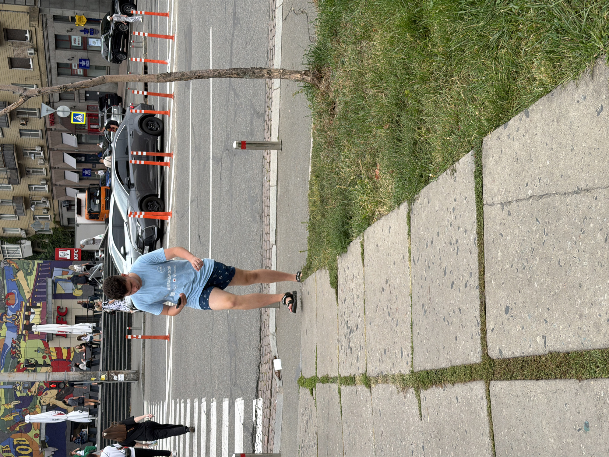 The image depicts a street scene in Kyiv, Ukraine. In the foreground, a young person wearing a light blue t-shirt and patterned shorts is walking across a pedestrian crossing, looking down at their mobile phone. They are wearing dark sandals. To the left of the pedestrian crossing, a portion of a sidewalk made of concrete slabs with grass growing between them is visible, bordering a grassy area. In the mid-ground, several cars are on the road, including a dark grey sedan and a lighter grey sedan. Orange and white bollards are placed along the road. In the background, buildings line the street, and a colorful mural is painted on the side of a building, featuring various figures and elements, including what appears to be a KFC restaurant. Other pedestrians are visible in the background, some walking and some sitting. A tree with a bare trunk stands to the right of the image. The overall environment suggests a daytime urban setting.