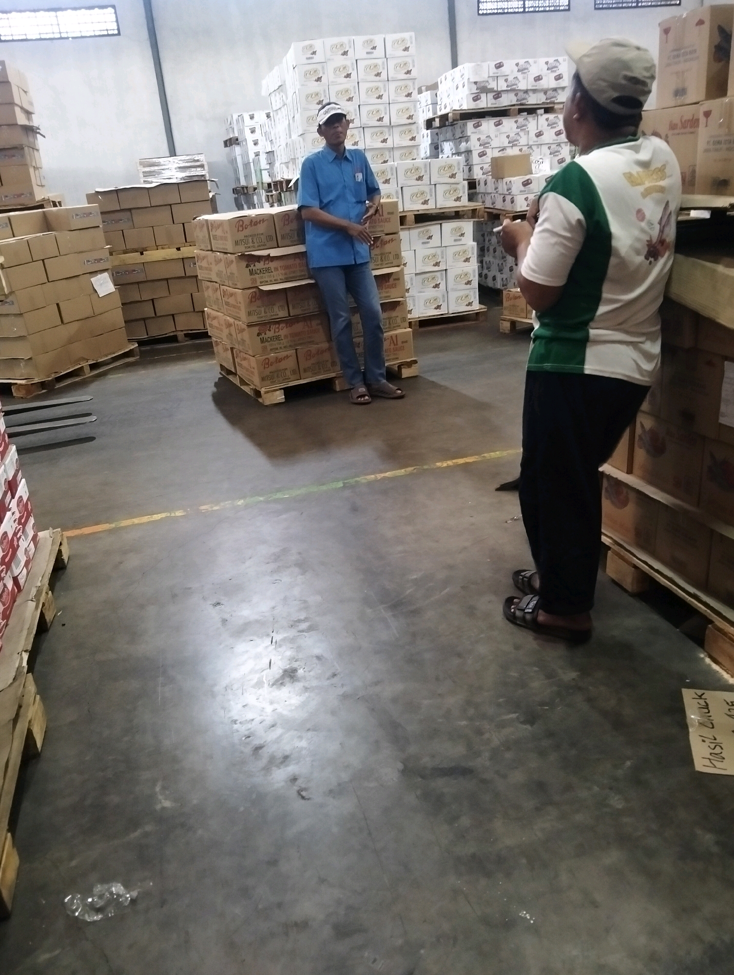 In Tanggulangin, Indonesia, two men are standing in a warehouse filled with stacked cardboard boxes. One man, wearing a blue shirt and a white baseball cap, stands near a pallet of boxes. The other man, wearing a white and green shirt and a beige baseball cap, stands with his back to the camera, holding a cigarette. The warehouse floor is concrete, with yellow and orange lines painted on it. Several pallets of boxes are visible throughout the space. One pallet has boxes labeled "Mackerel in Tomato Sauce." In the foreground, a piece of paper with handwriting on it is on the floor.