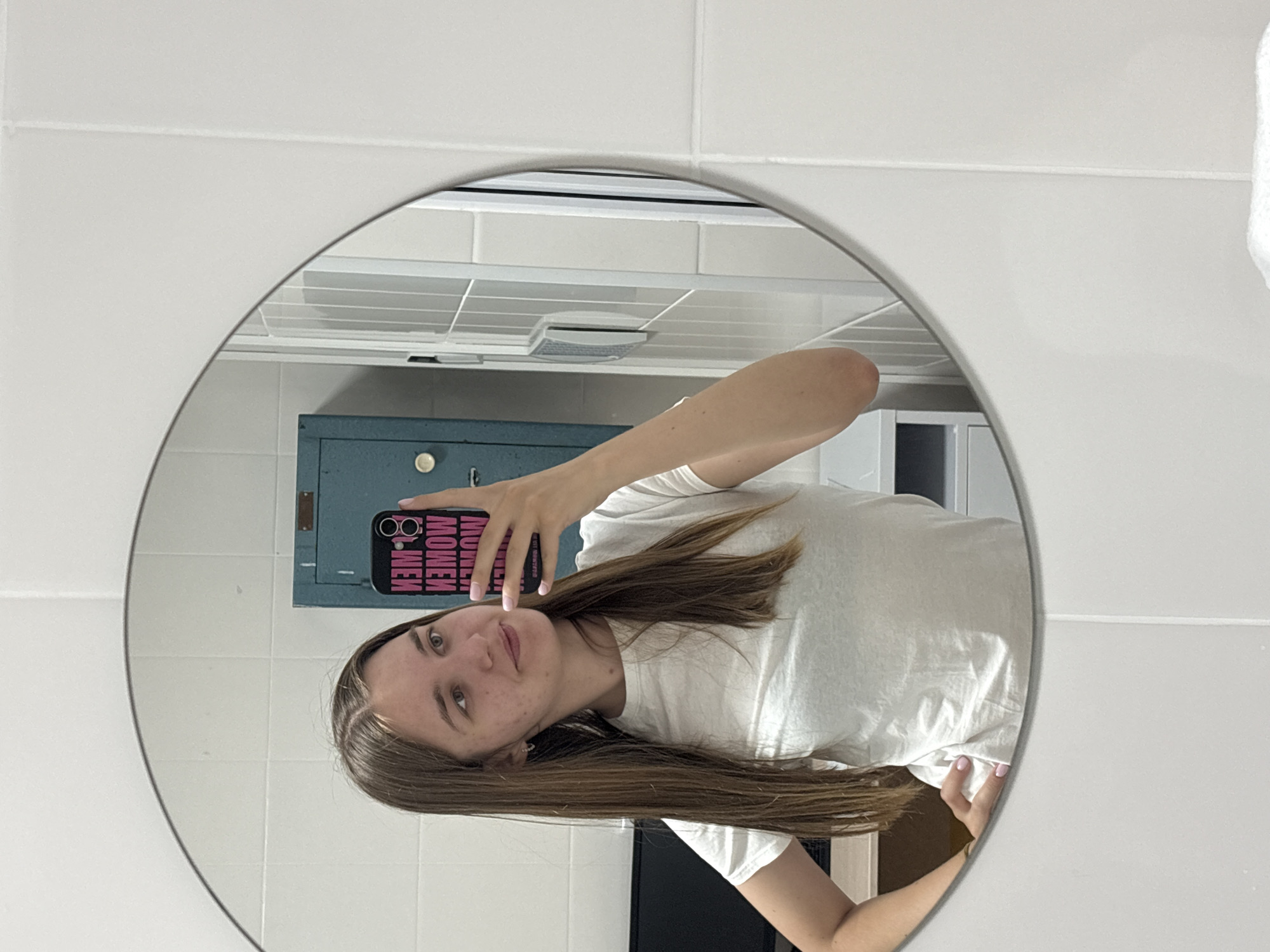 A young woman takes a selfie in a bathroom mirror. The background includes white tiled walls, a blue metal cabinet, and a ventilation system. The woman is holding a phone with a pink-lettered case. The location is Perm, Russia.
