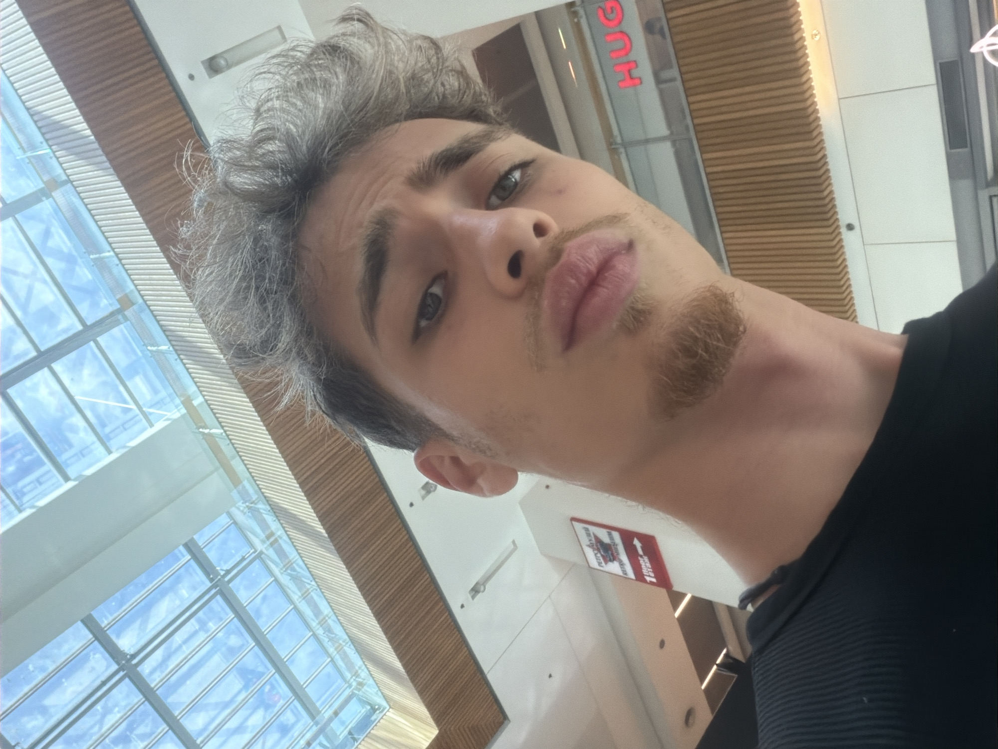 A close-up, low-angle selfie of a young man with fair skin, light brown hair with some grey strands, and a thin goatee. The man is wearing a black shirt. The background features a modern building interior with a skylight revealing a blue sky, and wooden slatted ceiling. A "HUGO" sign is visible. There's a sign in the background with "floor 1" on it. The man is likely in a shopping mall environment. The city of Varna, Bulgaria, cannot be determined based on the visible details in the image.