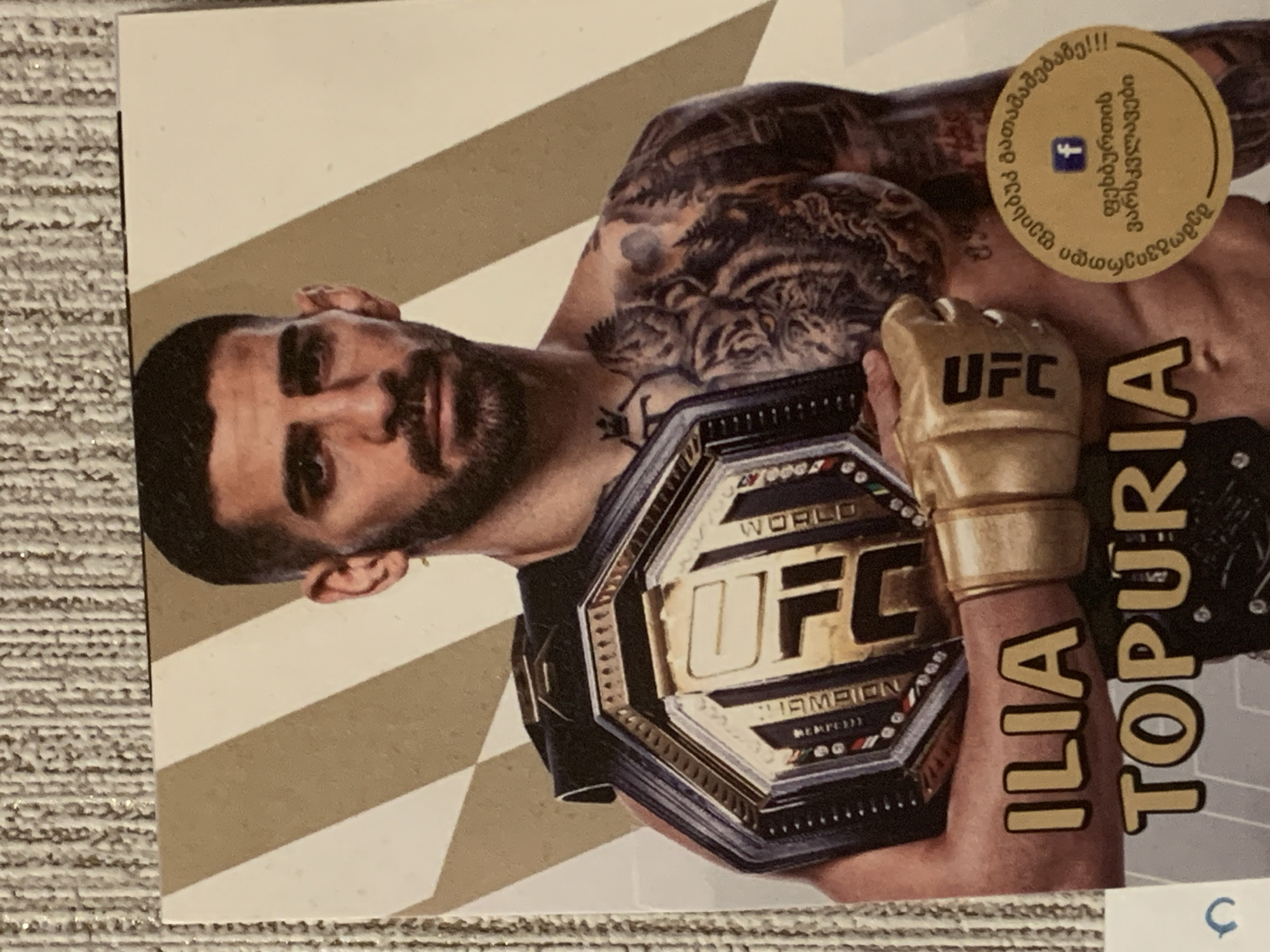 The image shows a card featuring a man holding a championship belt. The man is wearing a gold UFC glove on his left hand. He has dark hair, a beard, and tattoos on his chest and arm, including a tiger design. The championship belt is dark and gold with "UFC WORLD CHAMPION" written on it. The text "ILIA TOPURIA" is printed in large letters below the man. A circular sticker with Georgian text and a Facebook logo is visible on the man's right side. The background of the card has diagonal stripes in shades of gold and white. The card itself is placed on a textured surface, possibly a rug.