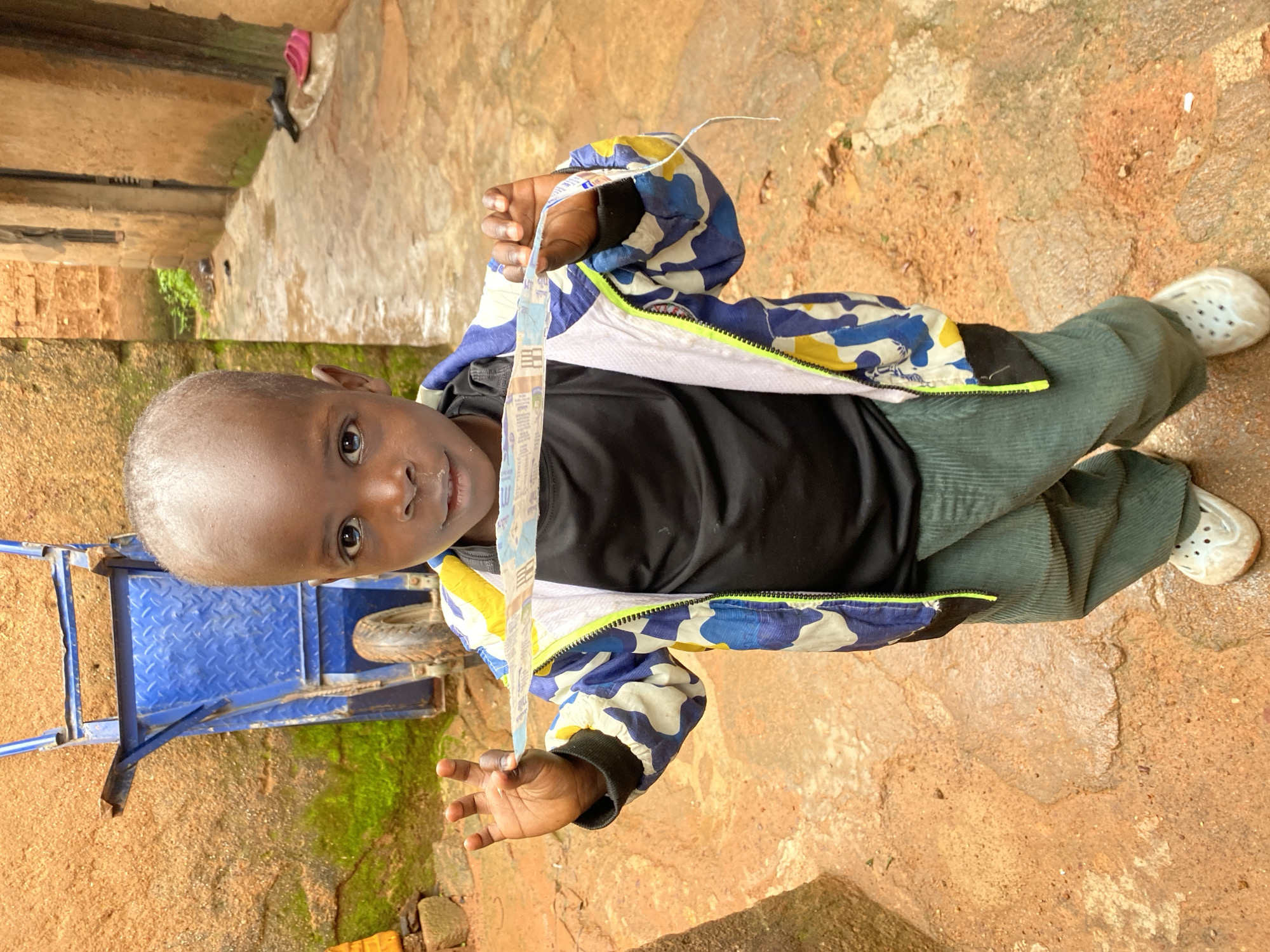 A young child, appearing to be of African descent with dark skin and a shaved head, stands outdoors in what seems to be a rural or semi-urban setting in Jos, Nigeria. The child is holding a torn strip of paper or fabric horizontally in front of them with both hands. They are wearing a black t-shirt, a jacket with a camouflage pattern of blue, white, and yellow, and green corduroy pants. White Crocs-style shoes are on their feet.
The background features a rough, orange-brown, uneven ground that appears wet in places, suggesting recent rain or damp conditions. To the left, a portion of a blue metal wheelbarrow is visible against a textured, reddish-brown wall covered in patches of green moss. To the right, a narrow alleyway or passage is lined with buildings constructed from what looks like mud or clay, with a concrete or paved walkway. A pair of pink flip-flops and black shoes are visible in the distance within the alley. The lighting is somewhat overcast, casting soft shadows. The overall impression is a candid, everyday scene in a possibly developing community.