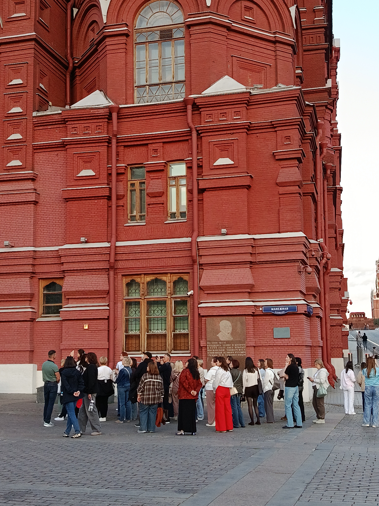 A group of approximately twenty to thirty people stands on a paved square in front of a distinctive red brick building. The building, characterized by its ornate architecture, features decorative white cornices, patterned brickwork, and various windows, including large arched ones on the upper floor and smaller, intricate ones with wooden frames and grilles on the lower levels. Vertical drainage pipes are visible along its facade.
The individuals in the foreground are casually dressed and appear to be listening or gathered as part of a tour, with most facing a common direction. The setting suggests late afternoon or early evening due to the soft lighting. The ground is paved with cobblestones, and a street or pathway with more people can be seen ascending in the background.
A blue street sign affixed to the building clearly reads "МАНЕЖНАЯ ПЛОЩАДЬ" (Manezhnaya Ploshchad), indicating Manezh Square. Below the sign, a memorial plaque with a bust is inscribed with Cyrillic text. The legible text on the plaque states: "НА ЭТОМ МЕСТЕ НАХОДИЛОСЬ ЗДАНИЕ, В КОТОРОМ В 1755 ГОДУ БЫЛ ОТКРЫТ МОСКОВСКИЙ УНИВЕРСИТЕТ ОСНОВАННЫЙ РУССКИМ УЧЕНЫМ М.В. ЛОМОНОСОВЫМ." This translates to: "On this place was located the building, in which in 1755 Moscow University was opened, founded by Russian scientist M.V. Lomonosov." The scene depicts a historical site with visitors engaging in sightseeing or a guided activity.