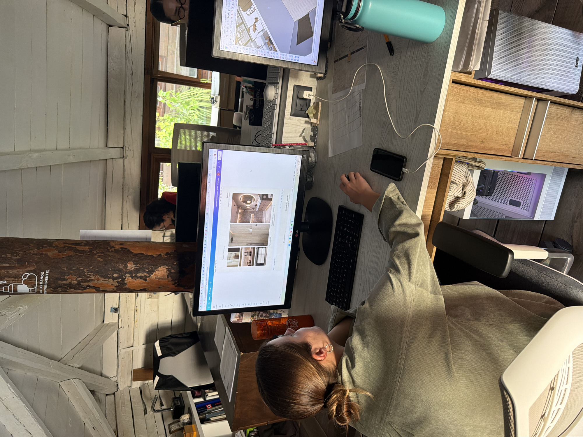 A person is seated at a desk with two computer monitors. The monitor in front displays a design with images of a bathroom interior. The other monitor shows a building plan. There is a cell phone plugged into a charger on the desk. A water bottle, keyboard, and documents are also on the desk. The person is wearing a green shirt and has their hair in a bun. Another person is visible in the background, near a window. The room has wooden beams and white walls, which suggests it may be an older building. A sign on a beam reads "LAVAR LAS MANOS CONSTANTEMENTE."