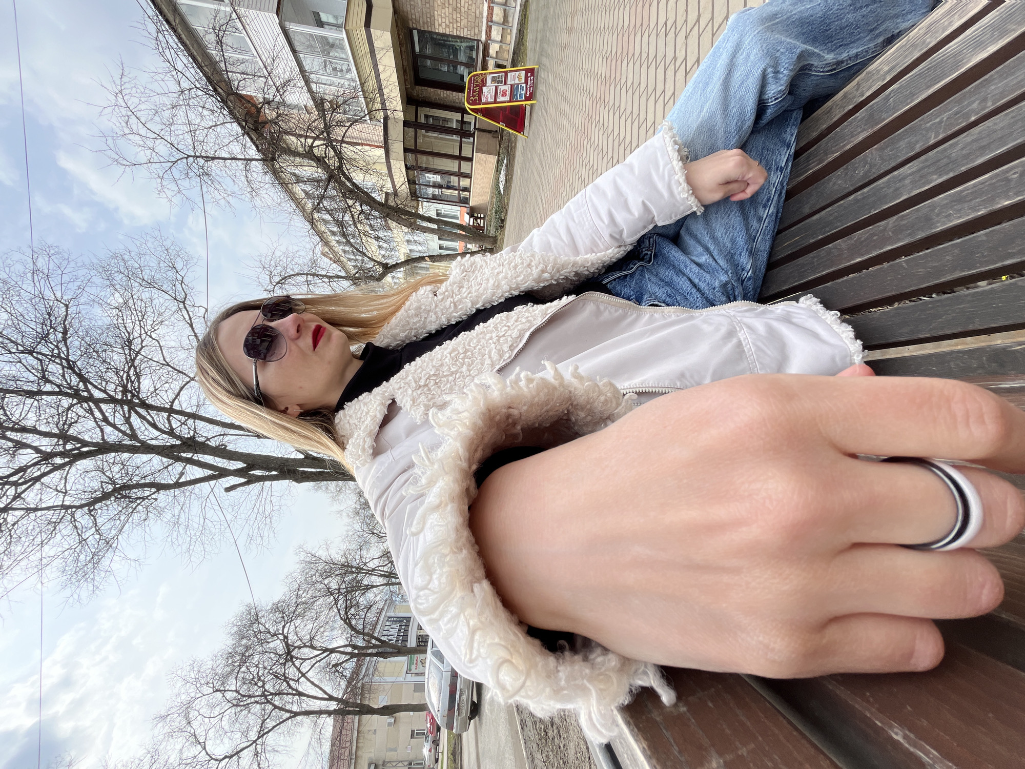 A medium, angled, outdoor shot shows a fair-skinned blonde woman reclining on a brown wooden bench, likely in Chaikovsky, Russia, given context clues. The woman wears black sunglasses, a white sherpa-collared jacket over a black turtleneck, and blue jeans. Her left hand, with a ring on the ring finger, is extended toward the camera. The bench is made of slats.
Behind her is a sidewalk made of interlocking pavers, followed by a building with a yellow sign. Bare trees are interspersed in the scene. The sky is cloudy. Cars are visible in the background. The image has an informal, selfie-like quality.