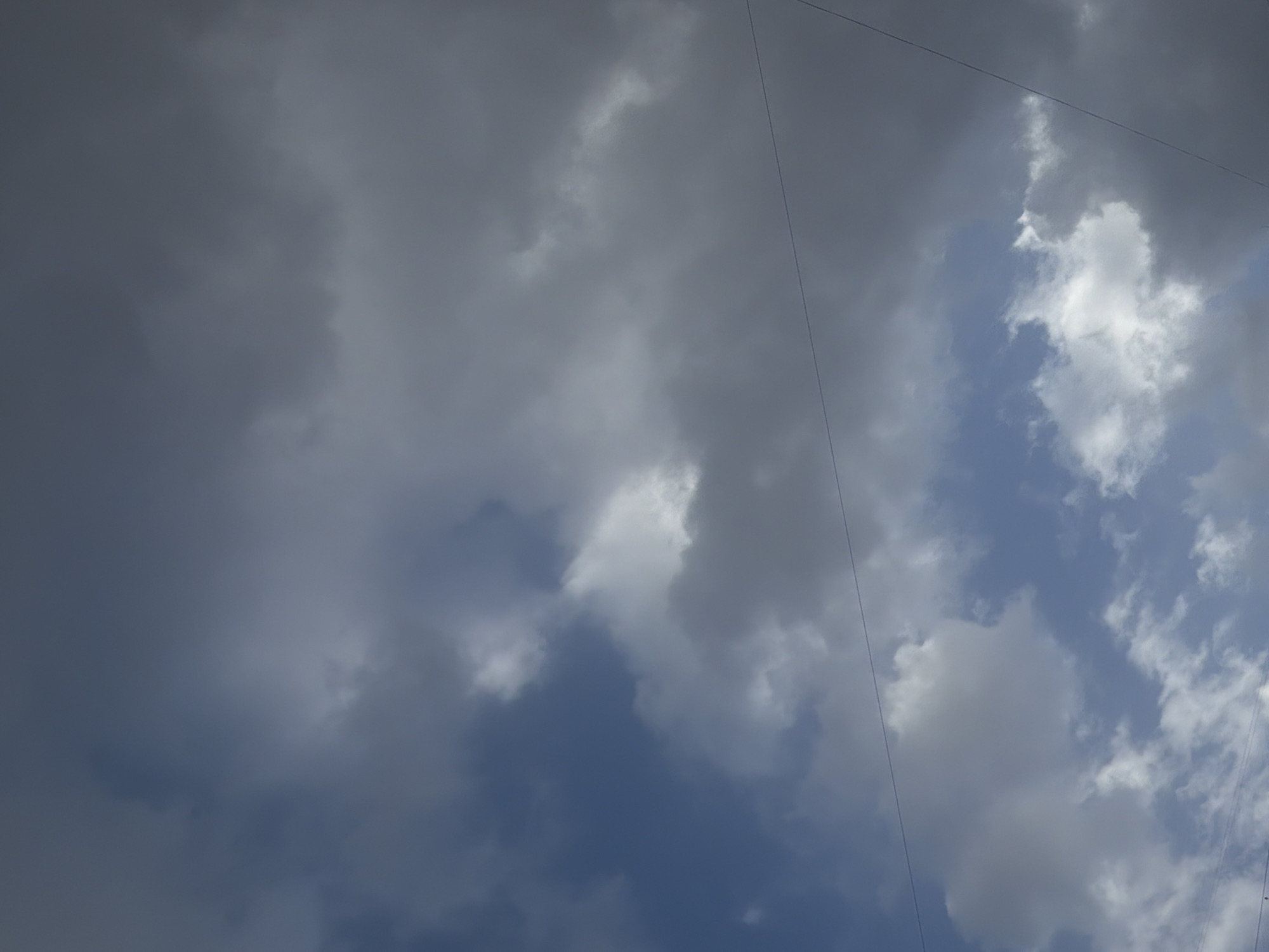 The image displays a dynamic sky dominated by clouds. The left side of the frame is covered by dense, dark grey clouds, indicating heavy cloud cover or an impending storm. Towards the center and right, the clouds become lighter and more scattered, revealing significant patches of clear blue sky. Some of the white clouds on the right exhibit bright, sunlit edges, contrasting with the darker, shadowed undersides of the heavier clouds.
Two thin, dark lines, resembling power lines or cables, traverse the sky. One runs diagonally from the upper left corner to the lower right, appearing to pass behind some of the clouds. Another parallel line is partially visible along the far right edge of the frame.
The overall impression is of a transitional weather condition, possibly a partially cloudy day or a sky clearing after or before rainfall in Perm, Russia. The time of day appears to be daytime, with sufficient sunlight to illuminate the clouds and reveal the blue sky. No specific activities, people, or text are visible.