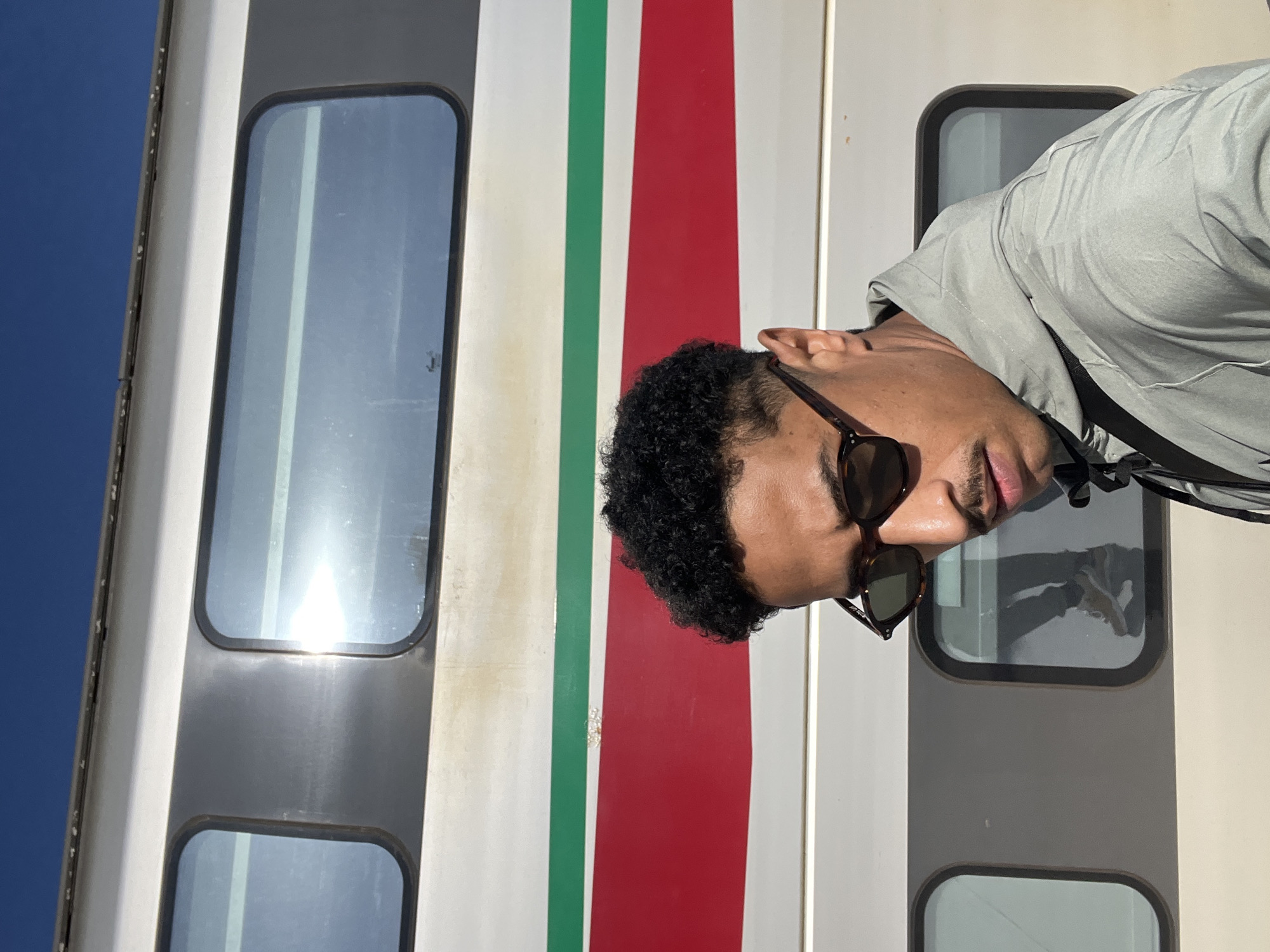 A young adult male is depicted standing outdoors on a sunny day, positioned in front of a stationary passenger train. The train, identifiable by its white body with prominent horizontal red and green stripes, indicates the location as a railway platform, consistent with the ONCF (Moroccan National Railways) in Tanger, Morocco.
The male subject has short, dark, curly hair with faded sides and a moustache. He wears dark, tortoiseshell-framed sunglasses and a light grey/silver hooded jacket with a slight sheen. A black strap is visible over his right shoulder. He is looking downwards and slightly to his right. The train's windows, with rounded corners, reflect a clear blue sky and strong sunlight. A reflection of another person's lower body, wearing light-colored sneakers and trousers, is visible in one of the train windows.