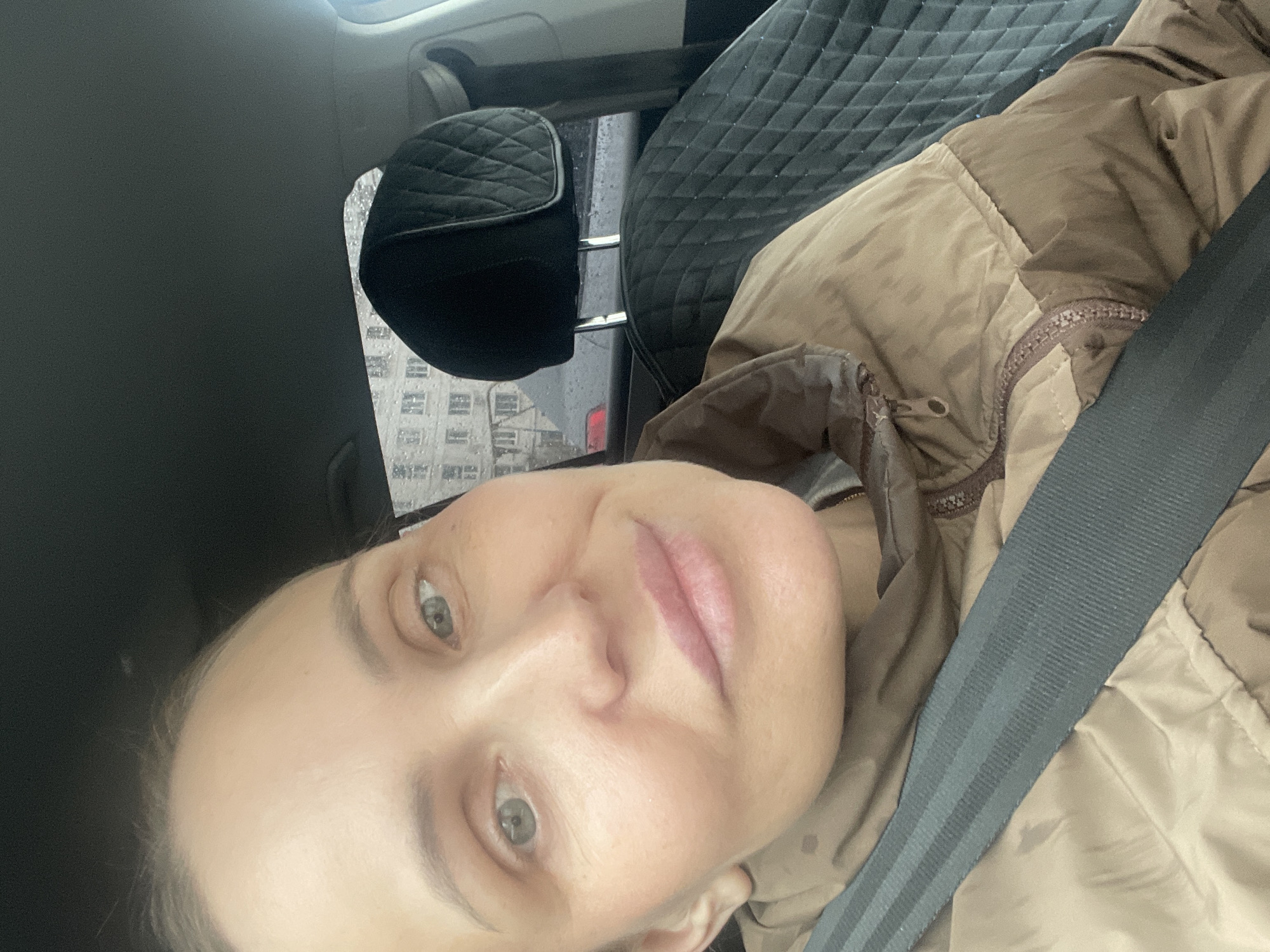This media file is a close-up, tilted selfie taken inside a vehicle. The primary subject is a person, likely a woman, with light-colored eyes, looking directly at the camera with a neutral expression. She is wearing a light brown or khaki puffer jacket with a zipper, and a dark grey seatbelt is visible across her chest.
The setting is the interior of a car. Details of the car include black, diamond-quilted seat upholstery and headrests. Through the car window, which is adorned with visible raindrops, an urban or suburban background featuring multi-story buildings, possibly apartment blocks, can be seen. The presence of raindrops suggests rainy or recently rainy weather, and the diffused lighting indicates an overcast day.
There is no visible text in the image. The general atmosphere is calm, and the person's expression does not convey strong emotions.
