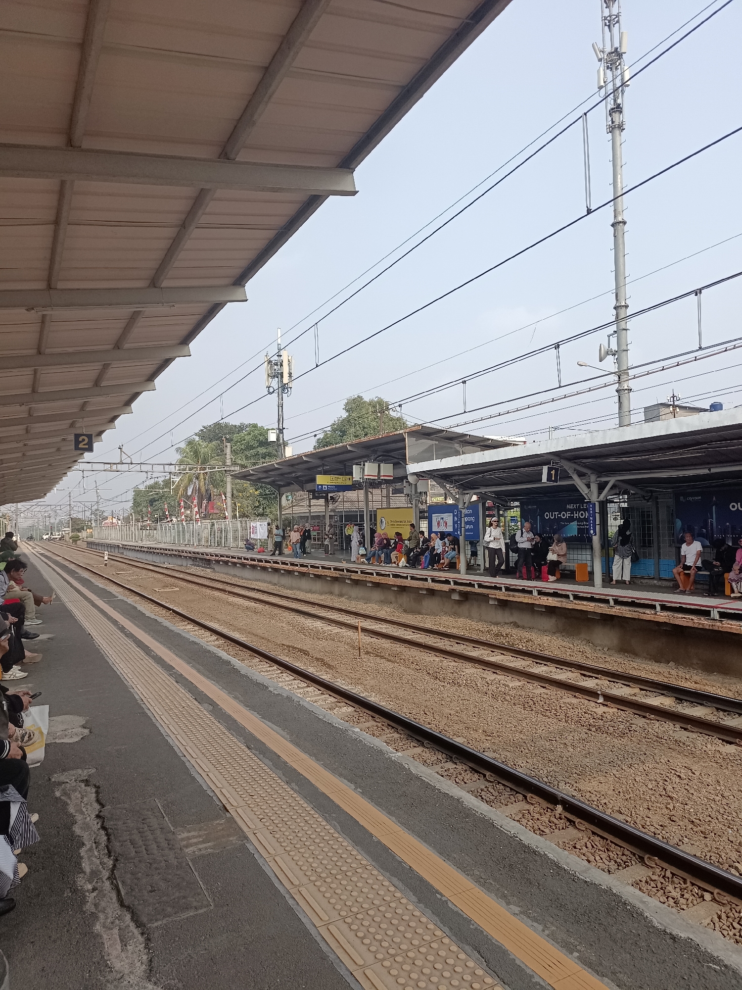 The image depicts a railway station platform in South Tangerang, Indonesia. On the platform, several people are waiting, some seated and others standing, with luggage visible. The platform is covered by a metal roof supported by pillars. Two sets of train tracks run alongside the platform, with ballast and gravel surrounding them. Overhead power lines and a cell tower are visible in the background. The scene is outdoors under an overcast sky.