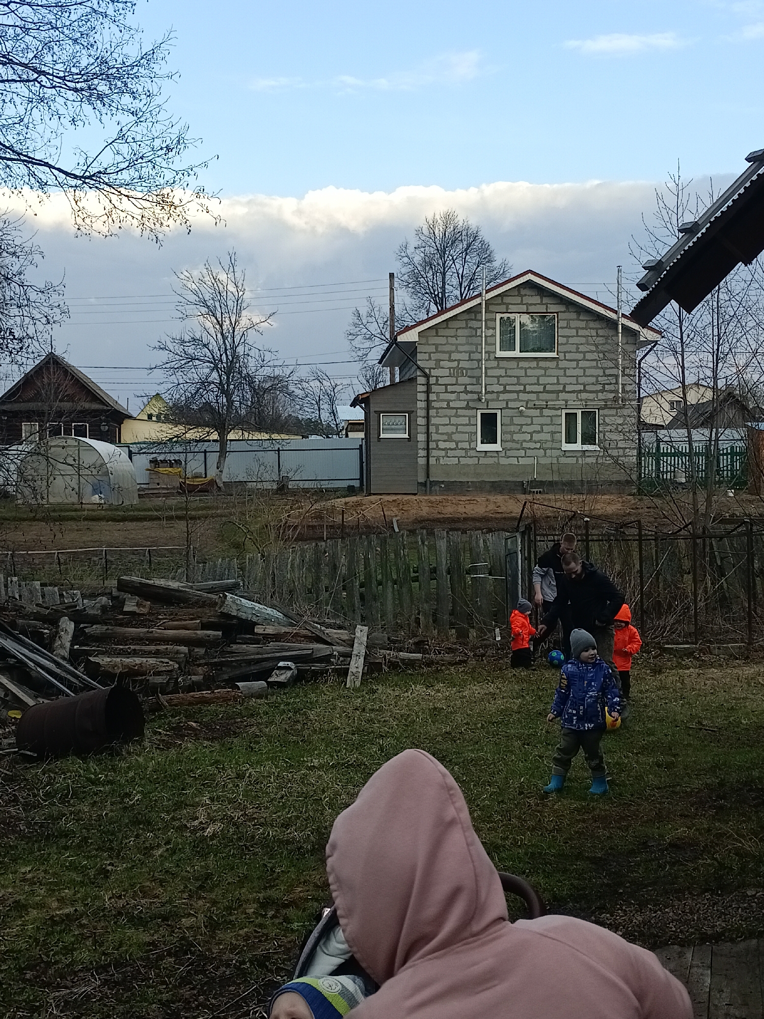 This image depicts an outdoor scene in Chaikovsky, Russia, likely a backyard or rural plot, involving several adults and young children. In the immediate foreground, the back of a person in a pink hoodie is visible, with a small child in a blue and white striped hat partially seen in front.
In the midground, an adult man in a black jacket is engaged with two young children, both wearing bright orange jackets, one of whom holds a blue ball. Another adult in a grey hoodie is close by. A third child, dressed in a blue patterned jacket, grey hat, and blue boots, stands slightly apart, holding a yellow toy. The ground is a mix of sparse grass and bare earth. To the left, a substantial pile of logs and an old metal barrel are visible.
The background is dominated by a two-story house built from grey cinder blocks, featuring white windows and a red roof, suggesting it is either under construction or recently completed, with scaffolding poles visible. To its left, a dark wooden house and a white tunnel greenhouse are present, all framed by a wooden fence. Bare trees, indicating either late autumn or early spring, are scattered throughout the scene. The sky is partly cloudy, with a distinct band of bright white clouds across the horizon and bluer sky above. The lighting suggests it is daytime, possibly late afternoon. No visible text is present. The overall atmosphere appears casual, depicting a family gathering or playtime.