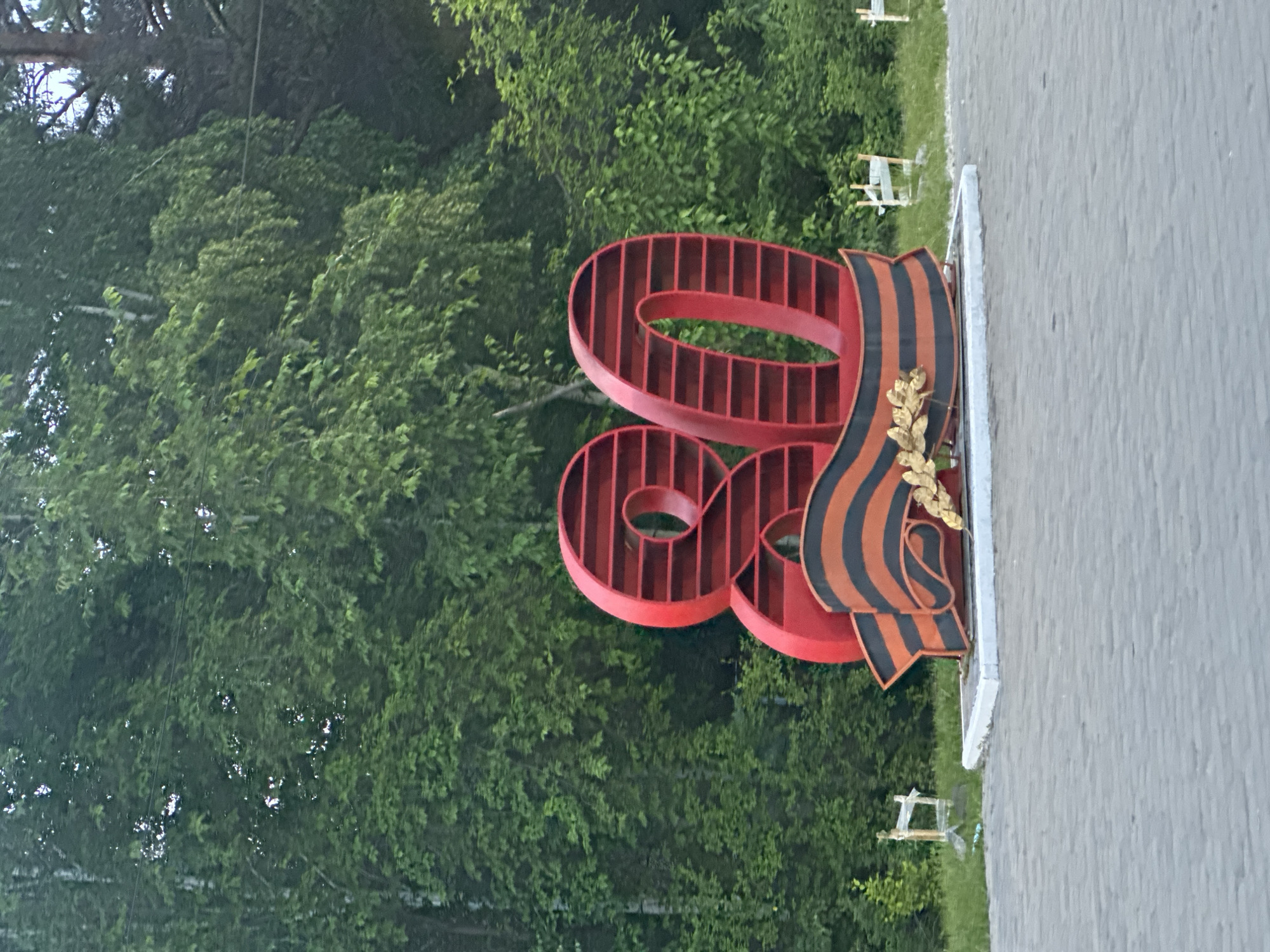 The image shows a large red metal sculpture of the number "80" in front of a backdrop of lush green trees. The sculpture is stylized with a ribbed design on the "8" and a St. George's ribbon with a laurel wreath attached to the right side of the "0". The St. George's ribbon is black and orange, a symbol commonly associated with Victory Day in Russia and other former Soviet republics.
The scene appears to be outdoors, possibly in a park or public space, given the presence of trees and a paved area to the right of the sculpture. In the background, glimpses of small white structures, perhaps benches or decorative elements, can be seen among the trees. The lighting suggests it might be daytime, with diffused light, and the overall atmosphere seems serene and commemorative. There is no visible text in the image. The sculpture's presence and the St. George's ribbon strongly suggest that the image is related to a celebration of an 80th anniversary, likely of a significant event or a veteran. Considering the location context of Shelekhov, Russia, this could be an commemoration of the 80th anniversary of the end of World War II or another important historical milestone.