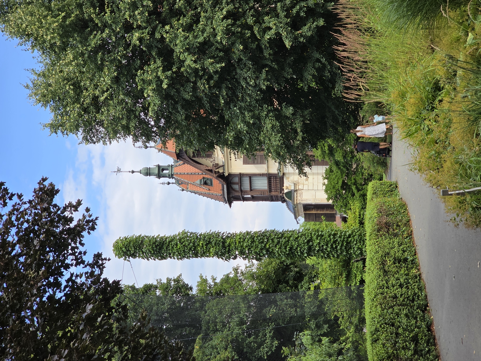 The image depicts a pathway through a lush green park with a large, ornate building in the background. A family consisting of two adults and a child are walking away from the camera down the pathway. One adult is pushing a stroller, and the child is holding the hand of the other adult. The building appears to be a historic villa with a red tile roof and intricate architectural details, including turrets and decorative windows. Tall, green foliage surrounds the pathway and the building, creating a serene and natural atmosphere. The sky is partly cloudy, indicating fair weather. The scene is set in the town of Zlín, Czechia, likely at the Vila Thomas Bata.
