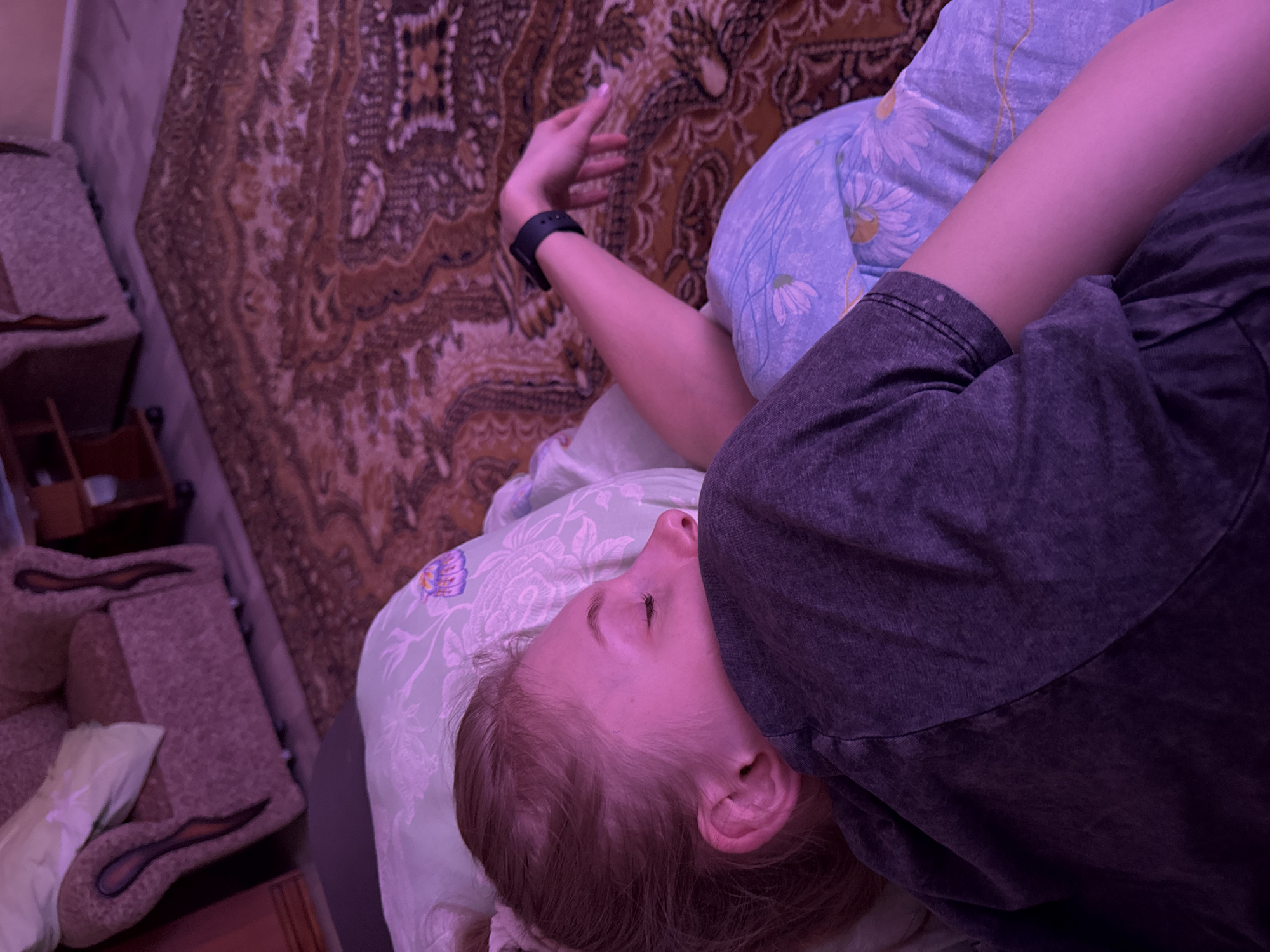 A fair-skinned young woman is lying on a bed in a room in Бородулино, Russia. The woman is lying on her back with her eyes closed and her head resting on a light green pillow with a floral pattern. She is wearing a dark gray t-shirt. Her right arm is extended above her head, and she is wearing a black smartwatch on her wrist. The bed has a patterned brown and beige bedspread. Next to the bed is a set of upholstered furniture. The room is lit with a purplish light.