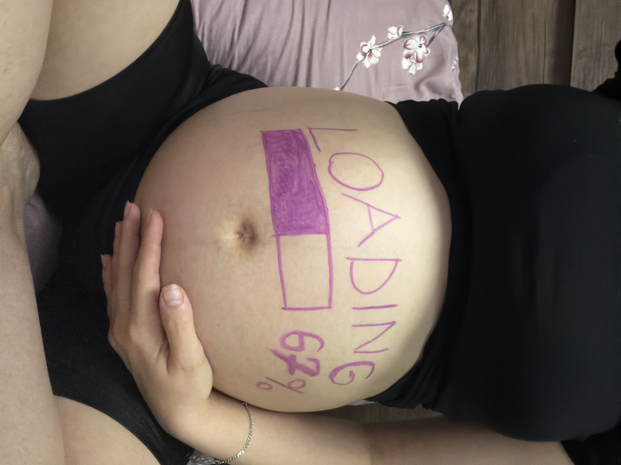A pregnant person's belly is the central focus of this image, with the word "LOADING" written in purple marker above a drawing of a progress bar. Next to the progress bar, "67%" is also written in purple. The person is wearing a black top and black shorts, and is resting on a pillow with a floral pattern. The background appears to be a wall. The image is taken from a close-up perspective, showing only the person's torso and hands. There is no information in the image that would indicate it is located in Tbilisi, Georgia.