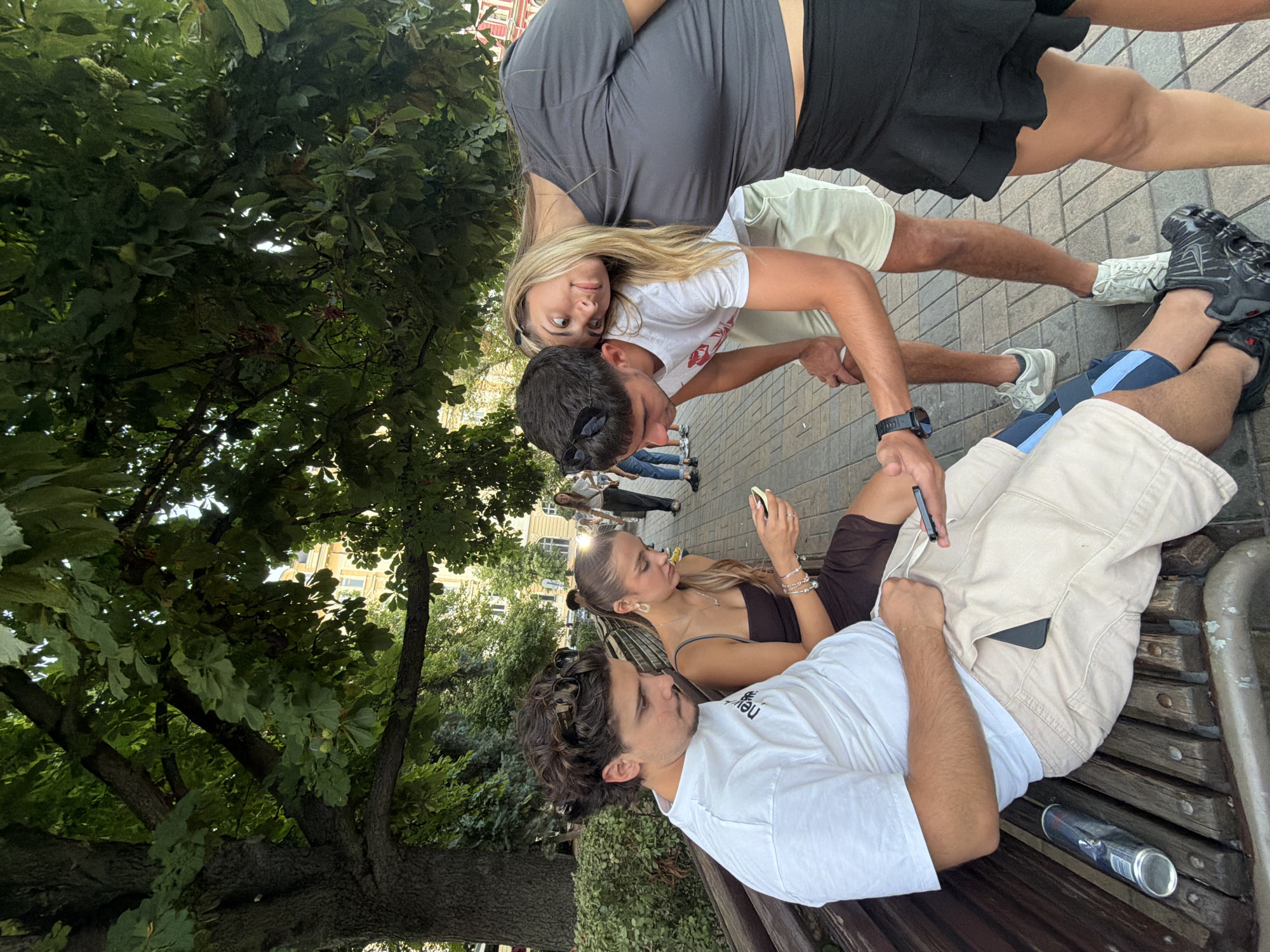 Four people are gathered on a park bench in Sofia, Bulgaria, under the shade of large trees. Two young men and two young women are present. The foreground features a man in a white t-shirt and beige shorts, holding a smartphone. Next to him, a young woman in a dark brown dress is also looking at her phone. Behind them, a young man in a white t-shirt and light shorts leans towards the woman to his right, his head resting on her shoulder. She is wearing a dark grey t-shirt and a black ruffled skirt. The background reveals a paved walkway and buildings, with other people walking in the distance. A crumpled metal can rests on the bench beside the man in the foreground. The overall atmosphere suggests a casual, relaxed outdoor gathering in an urban park setting.
