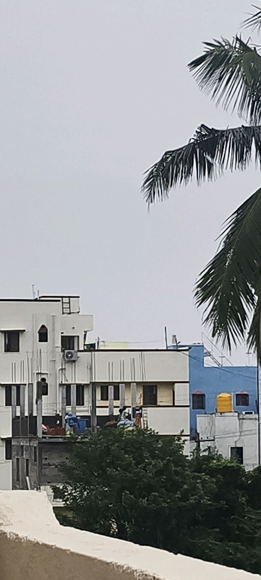 The image captures a construction site in Kundrathur, India, under an overcast sky. In the foreground, a white concrete ledge is visible. The mid-ground features a multi-story building under construction, with white and yellow exteriors and visible scaffolding. Several construction workers, dressed in blue, are present on the various levels of the structure, engaged in their tasks. An air conditioning unit is mounted on the side of one building. Adjacent to the construction is a blue-colored building with red-framed windows and yellow water tanks on its roof. A dense cluster of green trees partially obscures the lower levels of the buildings. Palm fronds from two palm trees are visible in the upper right portion of the frame, extending into the sky.