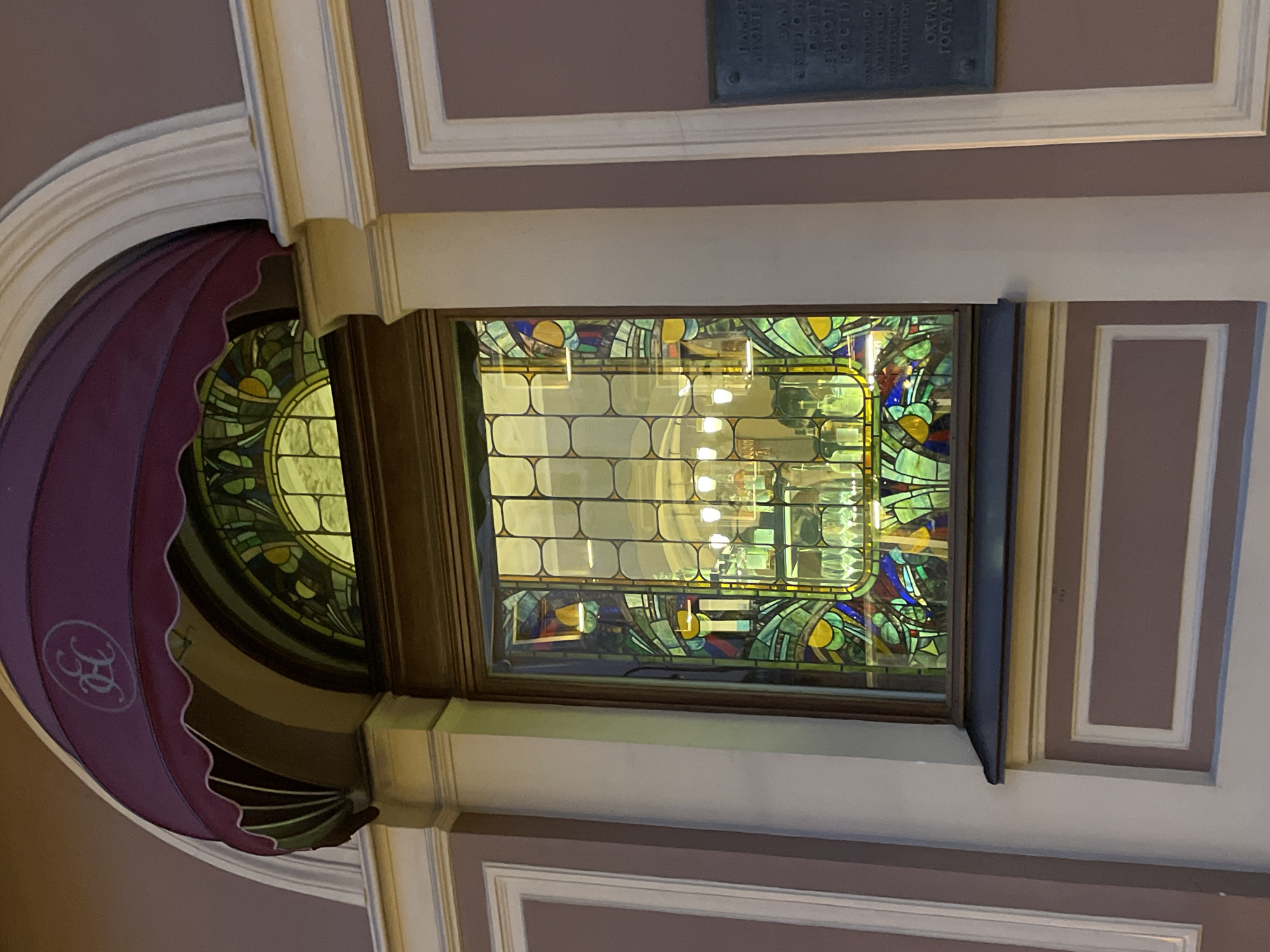 The image displays an ornate architectural detail, likely within a historical building in Saint Petersburg, Russia. The central feature is a rectangular stained-glass window, framed in dark wood. The window's design consists of a central grid of clear or translucent glass panes, reflecting bright interior lights, surrounded by colorful abstract patterns in greens, yellows, blues, and browns, indicative of an Art Nouveau or similar decorative style.
To the left, a large, semi-circular awning made of deep maroon or purple fabric is visible, featuring a scalloped edge. A white, stylized monogram, resembling two interlocking 'B' or 'R' letters, is prominent on the awning. This awning is situated beneath a curved white architectural molding.
The surrounding walls are a muted purplish-grey or taupe, embellished with elaborate white and light brown architectural trim and molding, contributing to a classical or historic aesthetic. Above the window, on the wall, a dark rectangular plaque is affixed. Though the text is largely unreadable in the main image, closer inspection of the crops reveals Cyrillic script. Partially discernible words like "ОХРАН..." (likely "protected") and "ГОСУДАРСТВ..." (likely "by the state") suggest it is a heritage plaque, designating the building as a protected cultural site.
The scene is well-lit by interior light sources, creating reflections in the glass. There are no people or discernible activities, emphasizing the static beauty of the architectural and decorative elements. The overall impression is one of elegance, historical richness, and meticulous preservation.