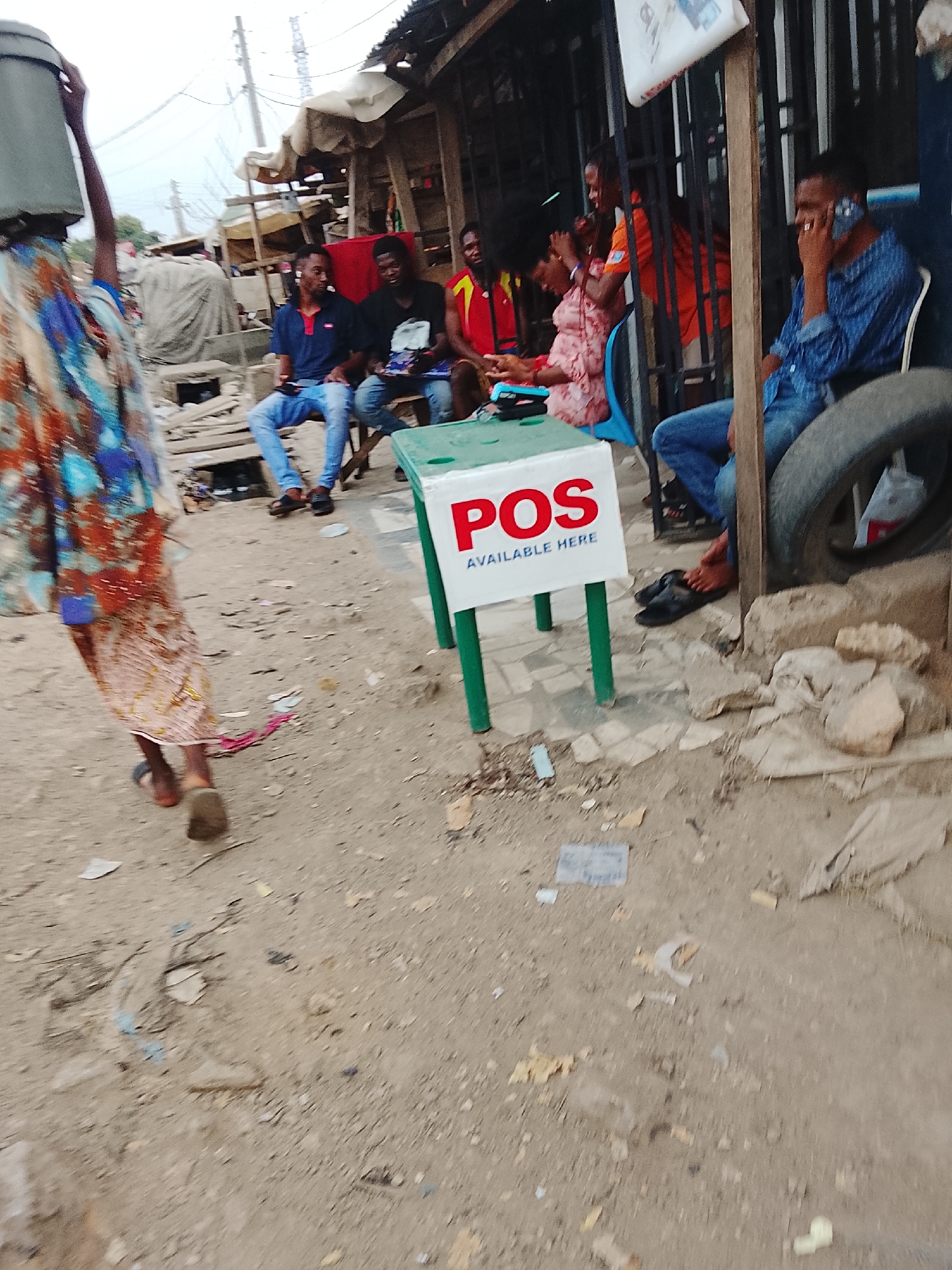 The image shows a street scene in what appears to be a lower-income area of Abuja, Nigeria. A woman is walking away from the camera, carrying a container on her head. In the background, several people are gathered, sitting and standing near a business. A sign on a small green table advertises "POS AVAILABLE HERE," indicating a point-of-sale service for electronic transactions. One man is on a mobile phone call. The environment is dusty and appears somewhat cluttered with various debris on the ground. The buildings in the background are simple and makeshift.