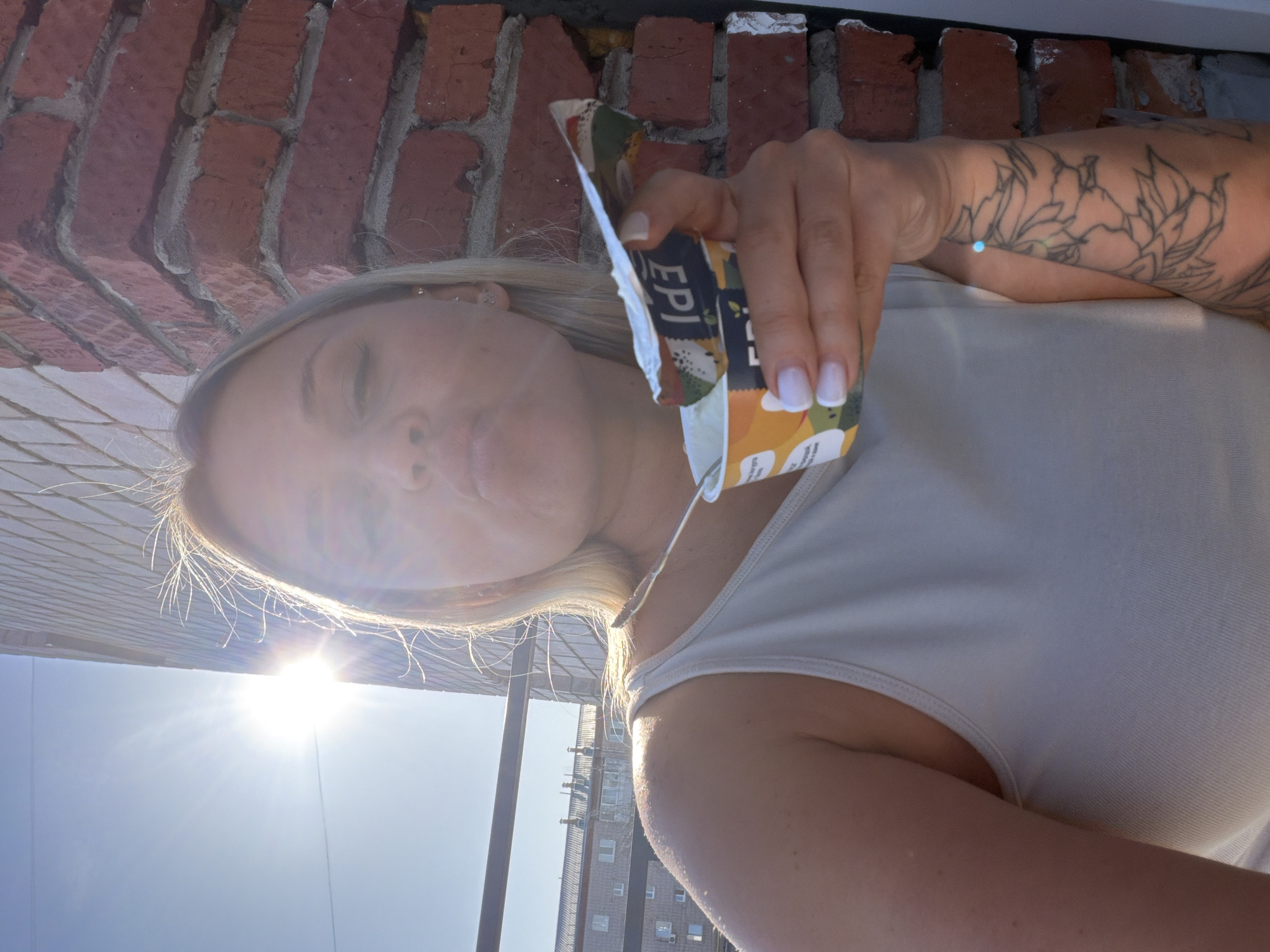 A woman with blonde hair is holding and consuming a food pouch outdoors. She is wearing a white tank top. She has tattoos on her left arm. Behind her, there is a brick wall and a building in the distance. The photo was taken in Sarapul, Russia. The sun is visible in the image.
