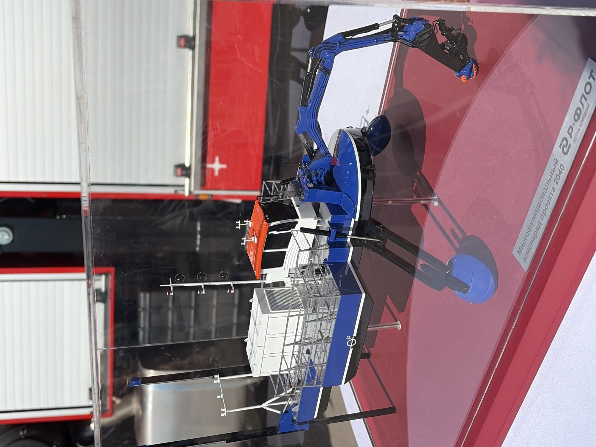 This is a detailed, forensic, matter-of-fact description of the image:
The image shows a model of a blue and white boat with an orange and white cabin and a blue excavator arm. The model is displayed on a red surface, enclosed in a clear plastic case. In the background, out of focus, is a large red object with white horizontal lines and a white cross, possibly part of a vehicle or structure. The model is supported by black legs. A small metallic plaque is visible at the bottom right of the red surface, with text that appears to be in Russian, indicating it is a model of a "Multifunctional Amphibious Dredger Project 2040" by "R-Pilot". There are no people or animals visible in the image. The lighting suggests an indoor display environment. The location is identified as the city of Bogorodsk, Russia, though this specific scene is not visually evident from the image itself, but is provided as context.