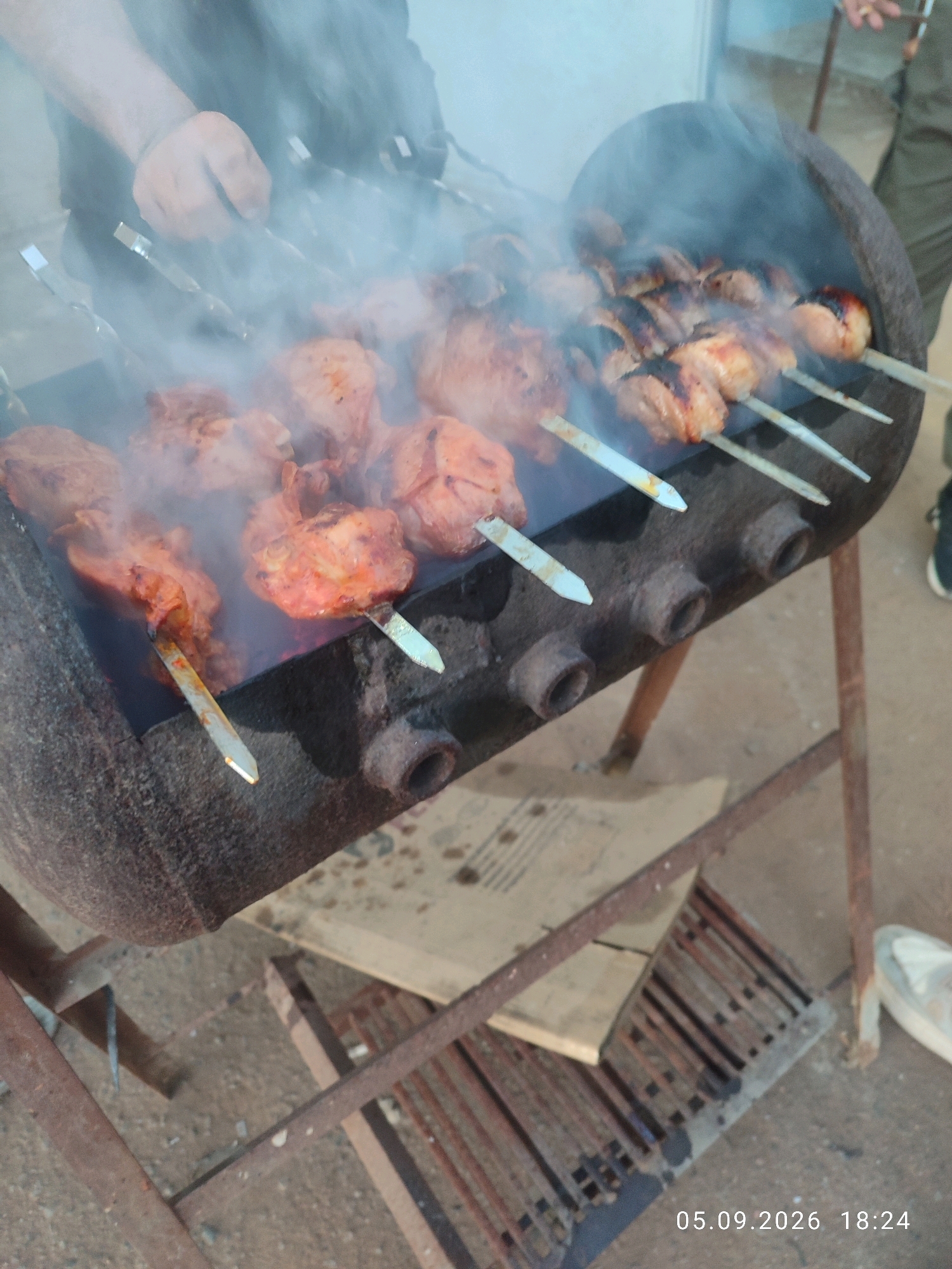 A horizontal, eye-level outdoor shot shows meat cooking on skewers over a makeshift grill. The grill appears to be constructed from a repurposed metal barrel, resting on a rusty metal stand. Smoke billows from the cooking meat, obscuring parts of the scene. Several skewers laden with pieces of raw and cooked meat are visible, some with a reddish hue, suggesting marination. A person's hands are partially visible in the upper left corner, near the skewers. The ground is a mix of dirt and gravel. In the lower right corner, a timestamp reads "05.09.2026 18:24".