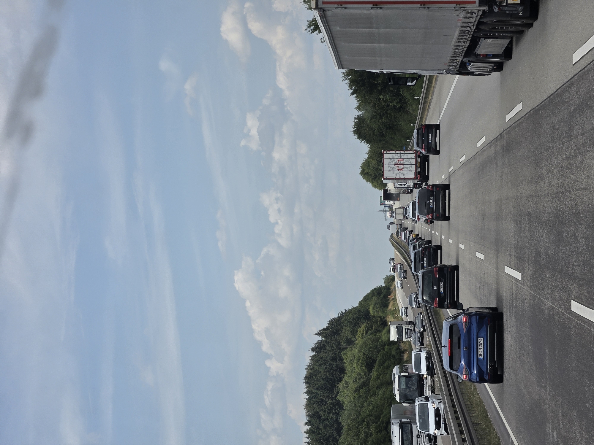 The image depicts a multi-lane highway in Oberselters, Germany, with moderate to heavy traffic. The road surface is asphalt, marked with white lane lines. The weather appears to be overcast with scattered clouds, and a patch of darker clouds is visible in the upper portion of the image.
On the highway, there are numerous vehicles including passenger cars, vans, and trucks. In the foreground, a blue hatchback is in the right lane, and a large semi-trailer truck is in the adjacent lane to its right. Further ahead, traffic is denser, with vehicles in all visible lanes. Several cars and trucks are distinguishable, some with visible license plates.
To the left of the highway, a dense line of evergreen trees borders the road, suggesting a rural or semi-rural setting. Beyond the trees, the sky transitions from a lighter blue near the horizon to darker gray clouds overhead. The overall impression is a typical highway scene during daylight hours. There are no visible people in the image.