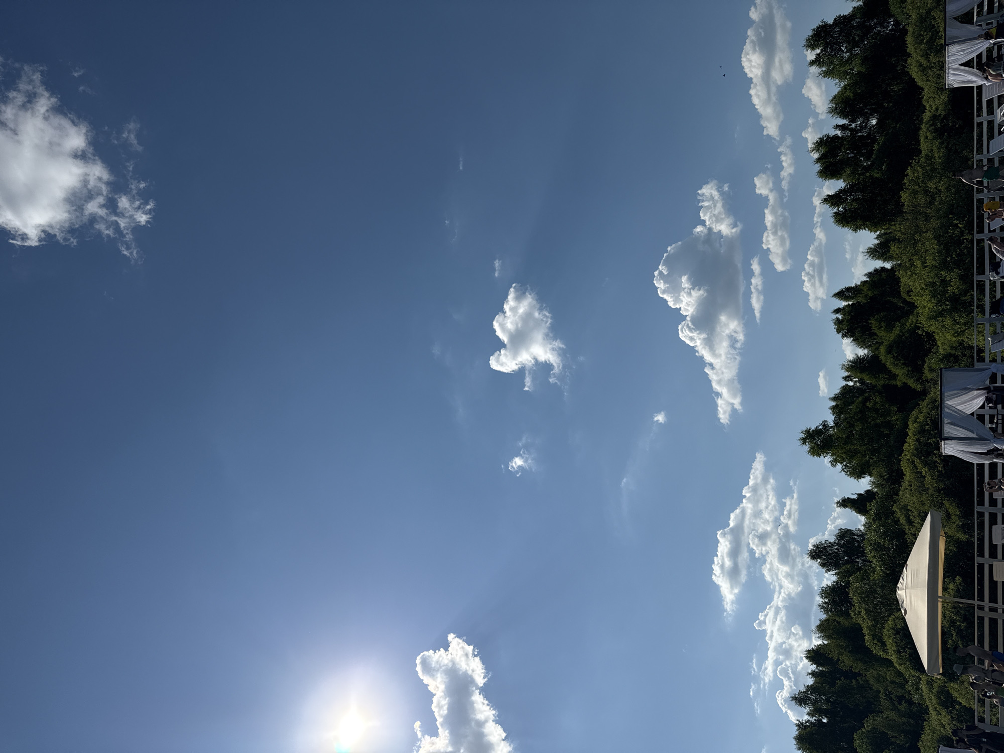 This image depicts a clear, bright day in Koloksha, Russia, under a vast blue sky dotted with fluffy white clouds. The sun shines intensely from the upper left corner, casting visible rays. In the foreground, a dense line of lush green trees forms a natural backdrop. Below the trees, a white railing delineates an area with lounge chairs and white canopies, suggesting a resort or recreational facility. A large, light-colored umbrella stands prominently near the center. Several people are visible in the lower portion of the image, some seated and others standing, enjoying the outdoor ambiance. The overall scene conveys a sense of a peaceful, sunny day in a natural setting within Koloksha.