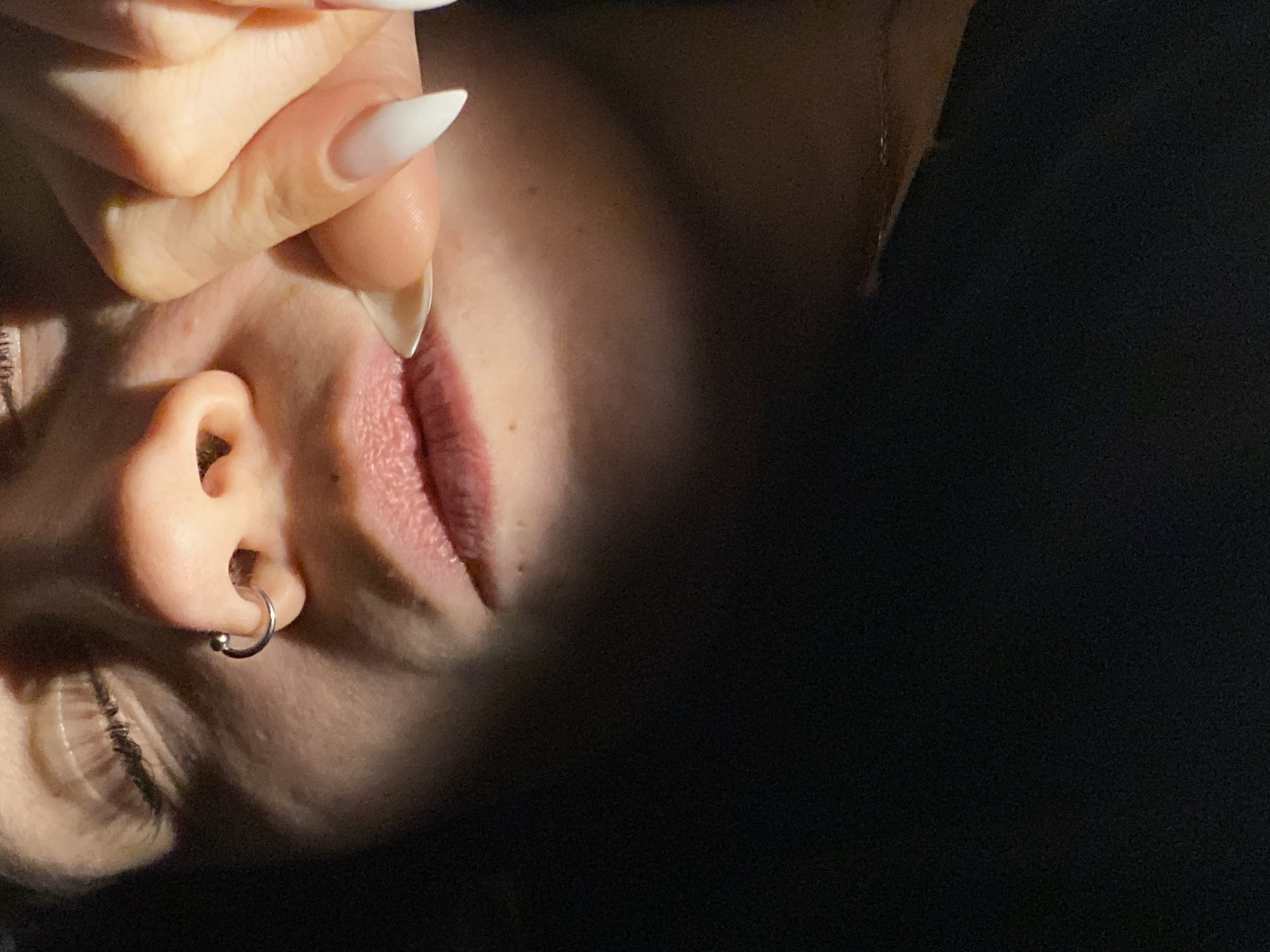 A close-up, tilted photograph depicts a person's face. The subject has fair skin, a lip piercing, and a silver earring. A hand with long, white, pointed fingernails touches the face near the mouth. The background is dark and undefined. The image lacks contextual details to place it in Chaikovsky, Russia, or any other specific location.
