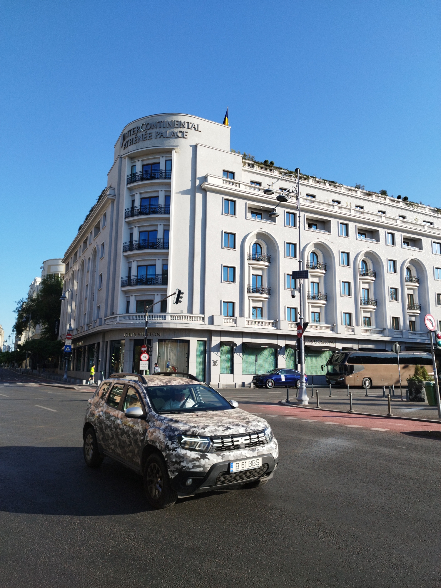 The image captures a street scene in Bucharest, Romania, likely in the vicinity of the InterContinental Athenee Palace Hotel, as indicated by the signage on the building. The hotel is a prominent, multi-story white building with arched windows and balconies. In the foreground, a Dacia Duster vehicle with a camouflage wrap is driving on the asphalt road. The vehicle's license plate reads "B 61 BBS". Several traffic lights and street lamps are visible. Further down the street, a brown bus and a blue sedan are also present. Pedestrians are visible on the sidewalk near the hotel, one of whom appears to be wearing a bright yellow vest. The sky is clear and blue, suggesting fair weather. The environment appears to be an urban setting with a mix of architectural styles.