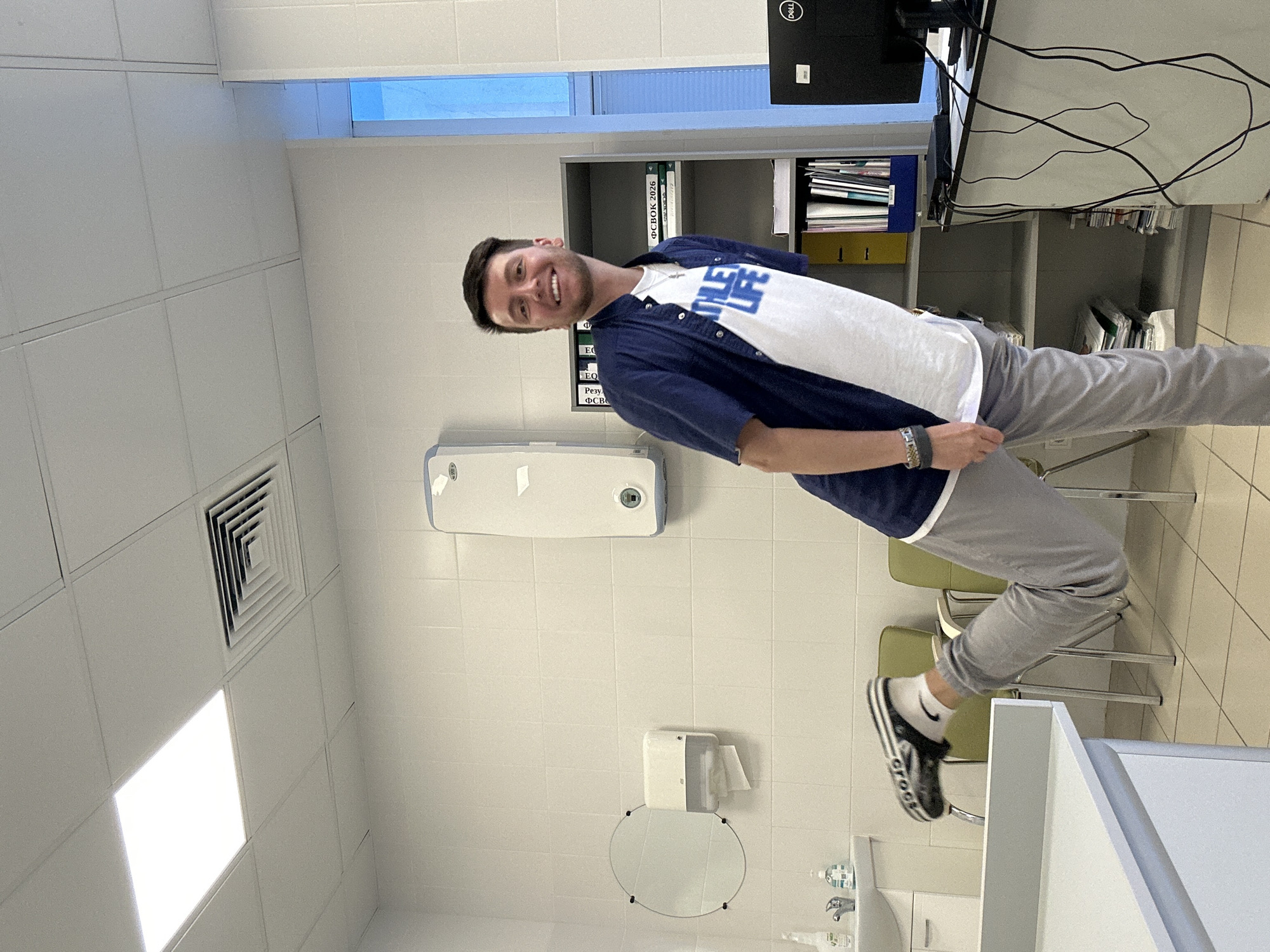 A man is captured in a vertical, full-body shot, smiling as he stands in what appears to be a well-lit, tiled room. He is wearing a white t-shirt with a blue graphic on it, a blue unbuttoned short-sleeved shirt over the t-shirt, grey sweatpants, and black and white striped Crocs with white socks. He has dark hair and appears to be in his late twenties or early thirties. His left hand is resting on his grey sweatpants, and he is wearing a dark bracelet on his left wrist.
The background suggests an indoor, possibly clinical or office setting. To the man's left, there is a white tiled wall with a large ventilation grate near the ceiling, and a bright fluorescent light panel. Further to the left, a white dispenser is mounted on the wall. In the background, there is a tall gray shelving unit filled with binders and papers. A black Dell monitor is visible on top of the shelving unit, with black cables trailing from it. There are also several green chairs in the background. To the man's right, there is a white dispenser with paper towels, a small circular mirror, and a sink with faucets and bottles of liquid.
The lighting is bright and even, suggesting it could be daytime or that the room is well-illuminated by artificial lights. There is a faint blue light visible in a strip near the ceiling in the upper part of the image, which could be natural light from a window or another light source. The overall impression is that of a clean, functional space. The man's smile and relaxed posture convey a positive and happy emotion. There is no discernible text in the image apart from what appears to be on the binders in the shelf and the branding on the monitor.