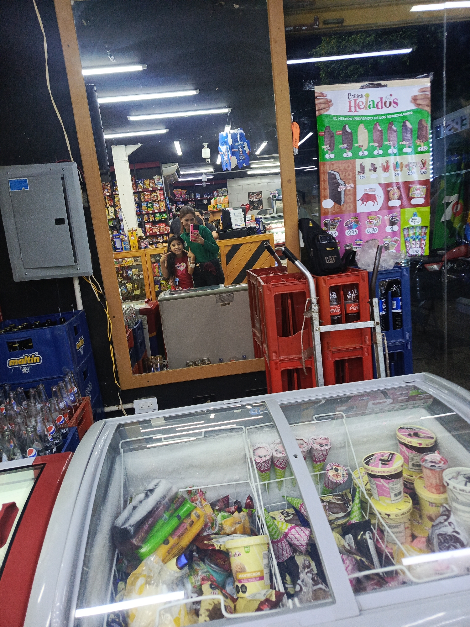 The image depicts the interior of a small retail space, likely a convenience store, in Altagracia, Venezuela. A freezer with a glass top filled with ice cream and frozen treats dominates the foreground. In the middle ground, there are stacks of beverage cases, including Maltin and Pepsi products. A red hand truck stacked with Coca-Cola bottles and beverage cases is present.
Reflected in a mirror, a woman and a young girl are visible. They appear to be taking a photo or selfie. The reflection also shows a glimpse of the store's interior, including shelving stocked with goods.
A promotional poster for "Crema Helados" is visible on the window to the right. The scene is illuminated by fluorescent lights and natural light from the exterior.
