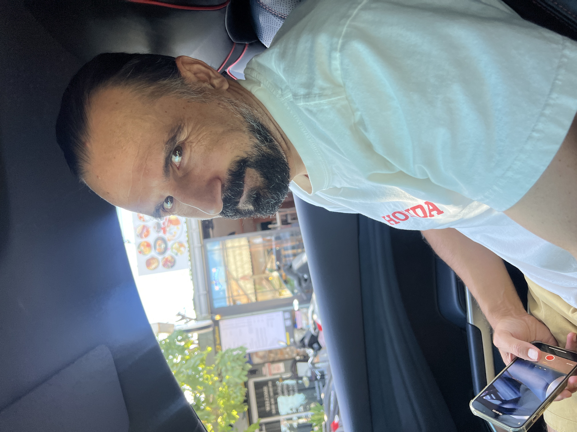 A man is taking a video inside of a car. The location outside the car has street signs for the "KENT & MILL ST WESTERVILLE" and a restaurant called "HOOTS AMERICAN GRILL".