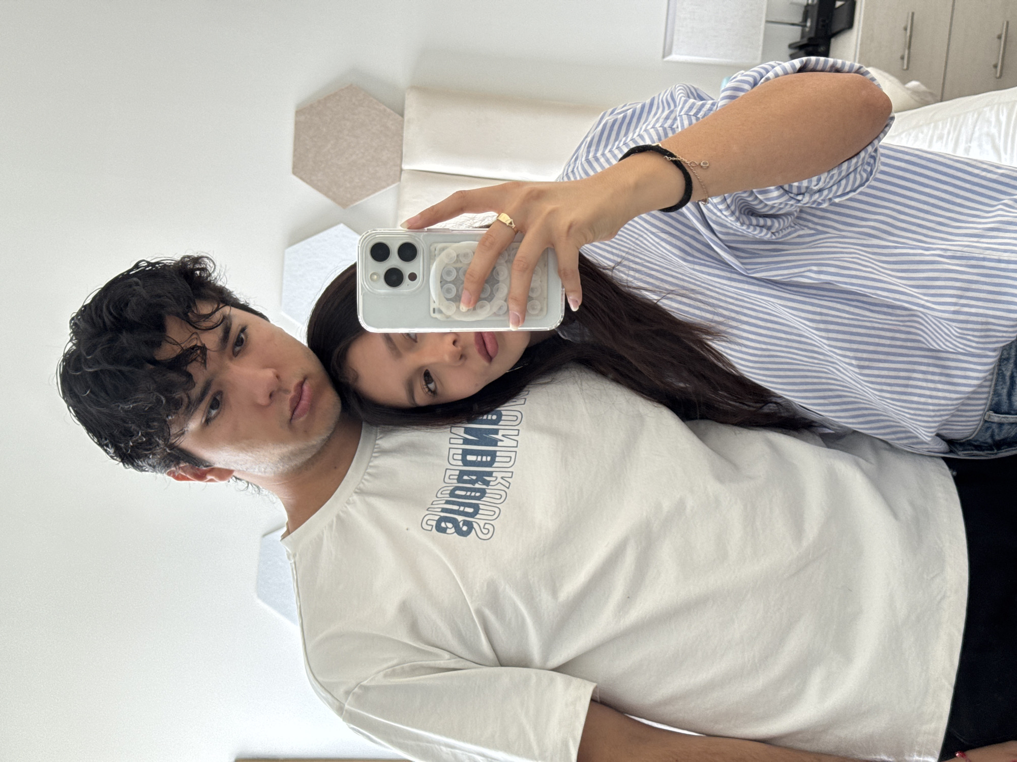 A young couple is taking a selfie indoors. The man has dark, curly hair and wears a white t-shirt. The woman, with long, dark hair, rests her head on his shoulder while holding a smartphone up to take the picture. She wears a striped blue shirt. The background includes a white wall with decorative hexagonal panels, a bed with white bedding, and a partially visible dresser with handles. The location is potentially San Francisco Ocotlán (Ocotlán), Mexico.