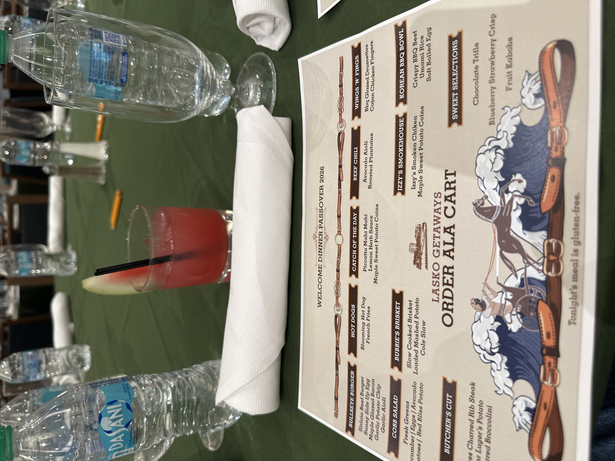 The image shows a table setting at what appears to be a welcome dinner for Passover 2026. The location is inferred from the menu titled "Lasko Getaways" which could be at a resort location in Aventura, United States of America, given the resort-style theme of the menu. A menu card stands prominently, listing various food and drink options in an "a la carte" style. Beverages, including bottled water, and a red-colored drink with a watermelon garnish are visible, indicating a meal or event in progress. The setting includes folded white napkins and potentially other guests at the table, though they are out of focus. The overall atmosphere is that of a celebratory or special occasion dinner. The menu indicates that tonight's meal is gluten-free.