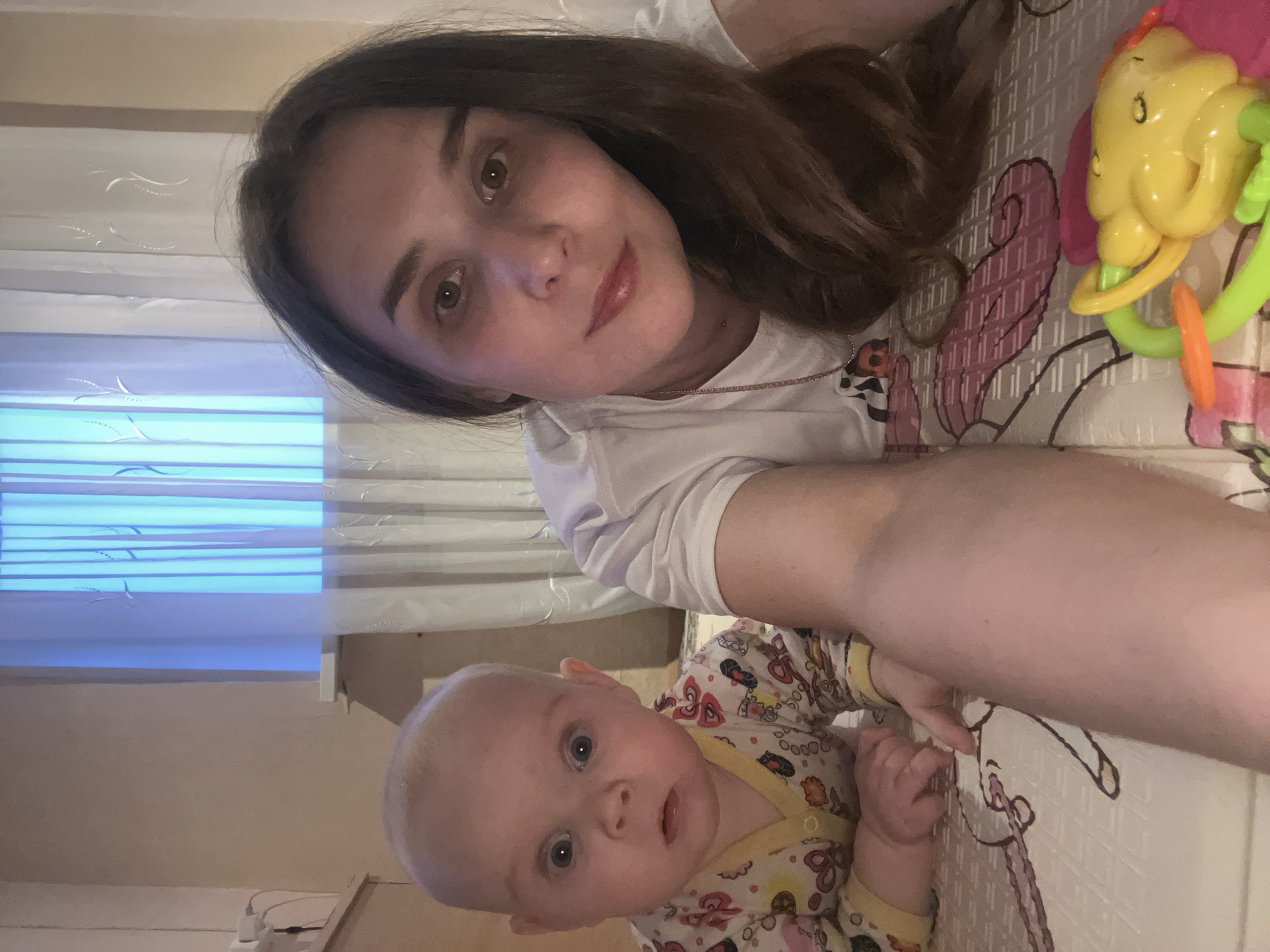 A woman and a baby are lying on a patterned surface. The woman has brown hair and is wearing a white shirt. The baby is wearing a patterned outfit with yellow trim. A yellow and green baby toy is next to the woman. The location is not discernible from the image.